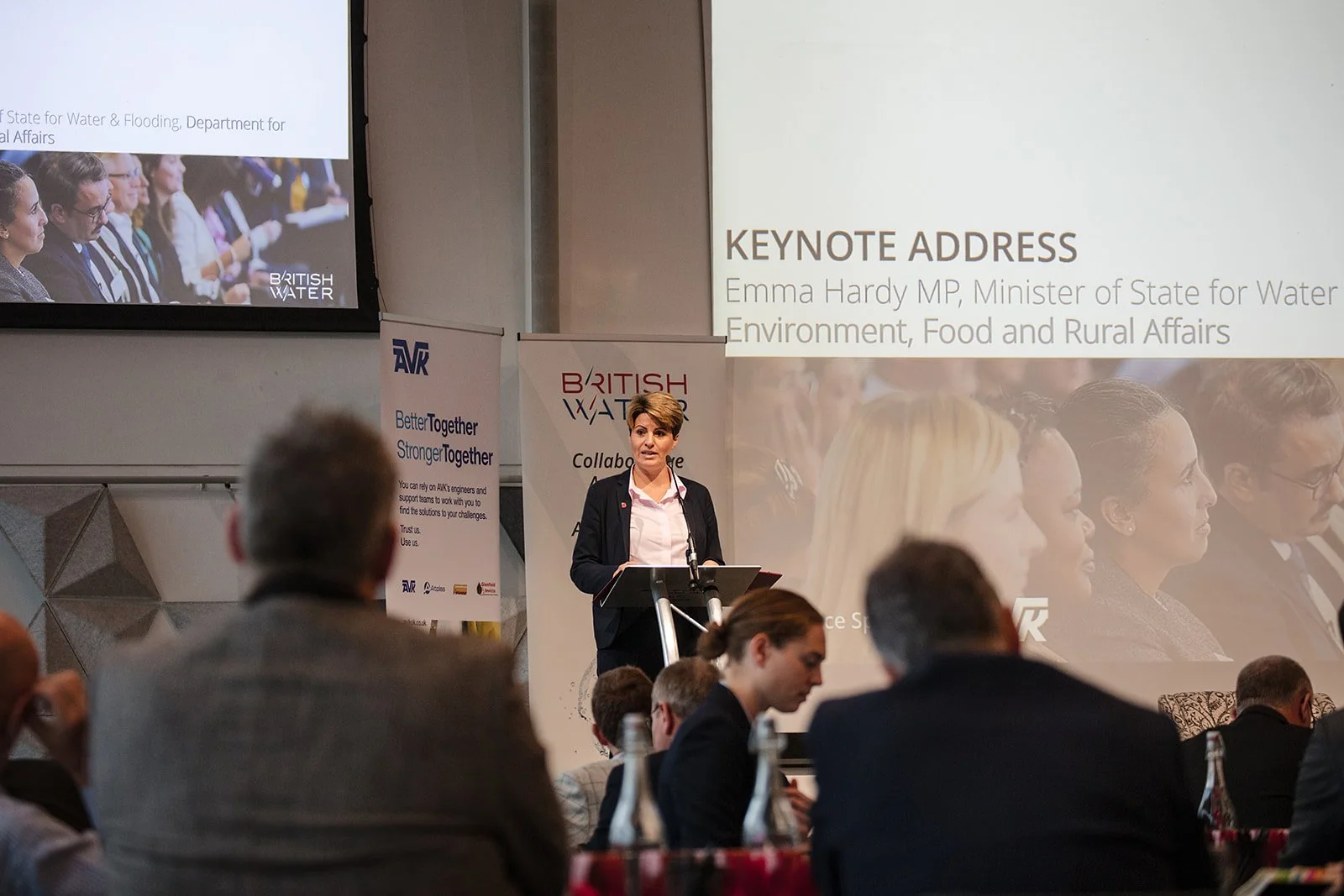 Emma Hardy MP giving a keynote address at a conference on water, environment, food, and rural affairs, standing at a lectern with a microphone, audience members seated in front of her, large screen behind displaying her name and title, and banners wi