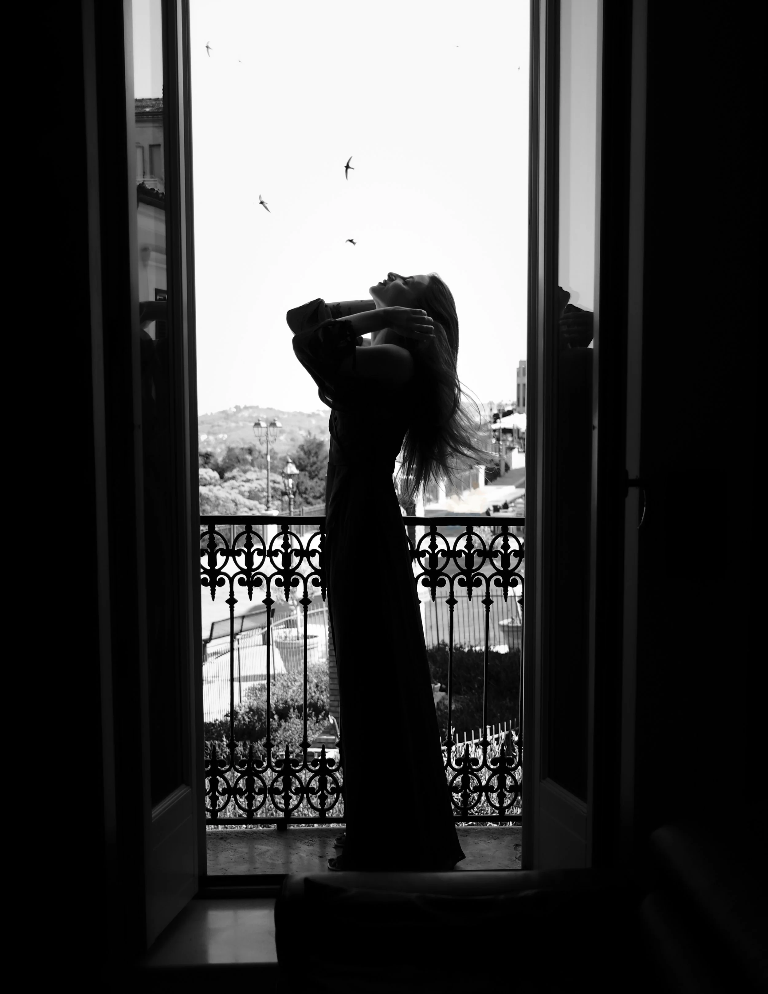Silhouette di una donna su un balcone, vista tramite una porta di casa, con uccelli che volano nel cielo.