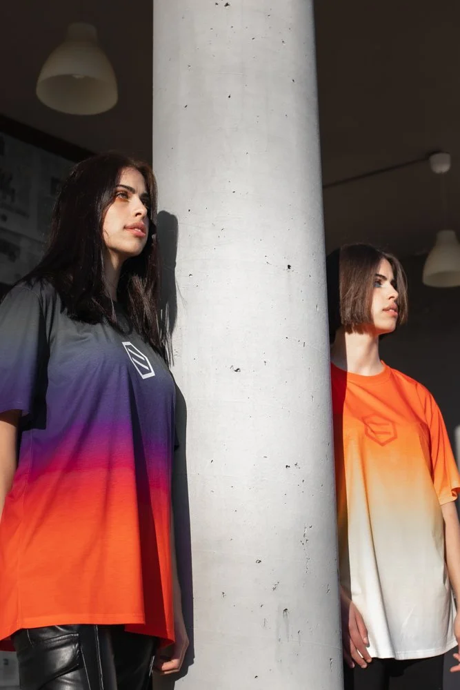 Due donne con magliette colorate, una con maglietta arancione e l'altra con maglietta blu e viola, stanno appoggiate a un pilastro bianco in un ambiente interno.