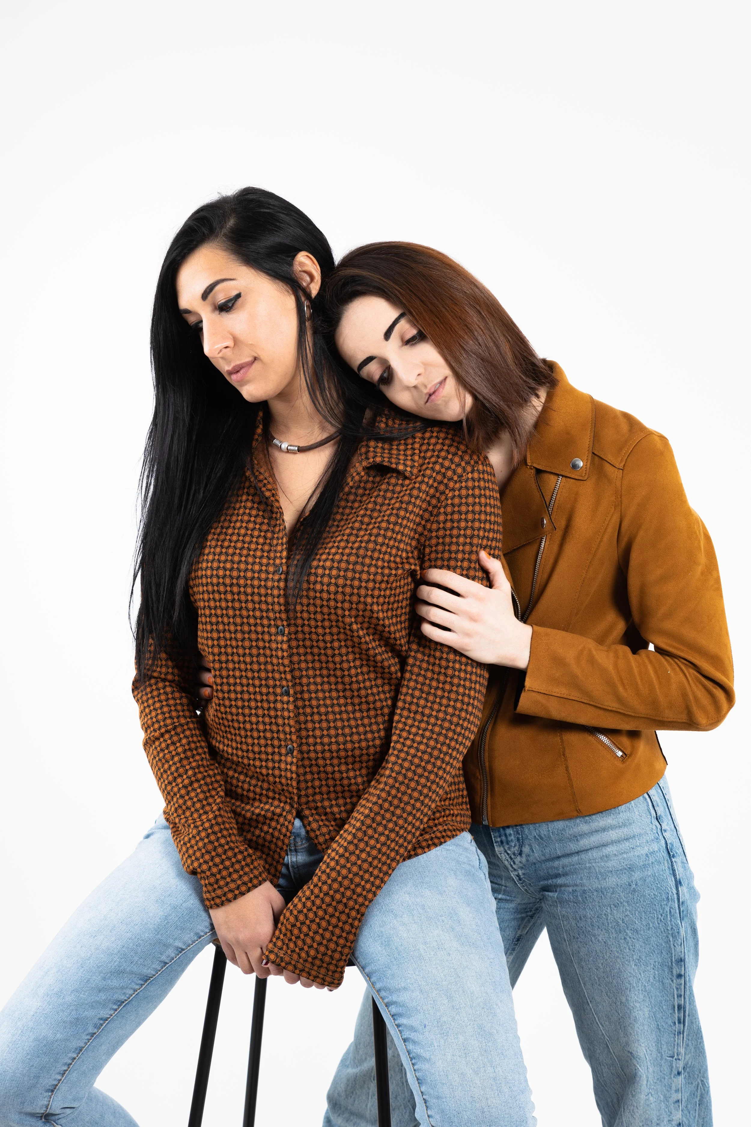 Due donne che mostrano empatia e conforto, con una che si appoggia all'altra, entrambe vestite casual con jeans e giacche marroni e nere, in un ambiente bianco senza sfondo.