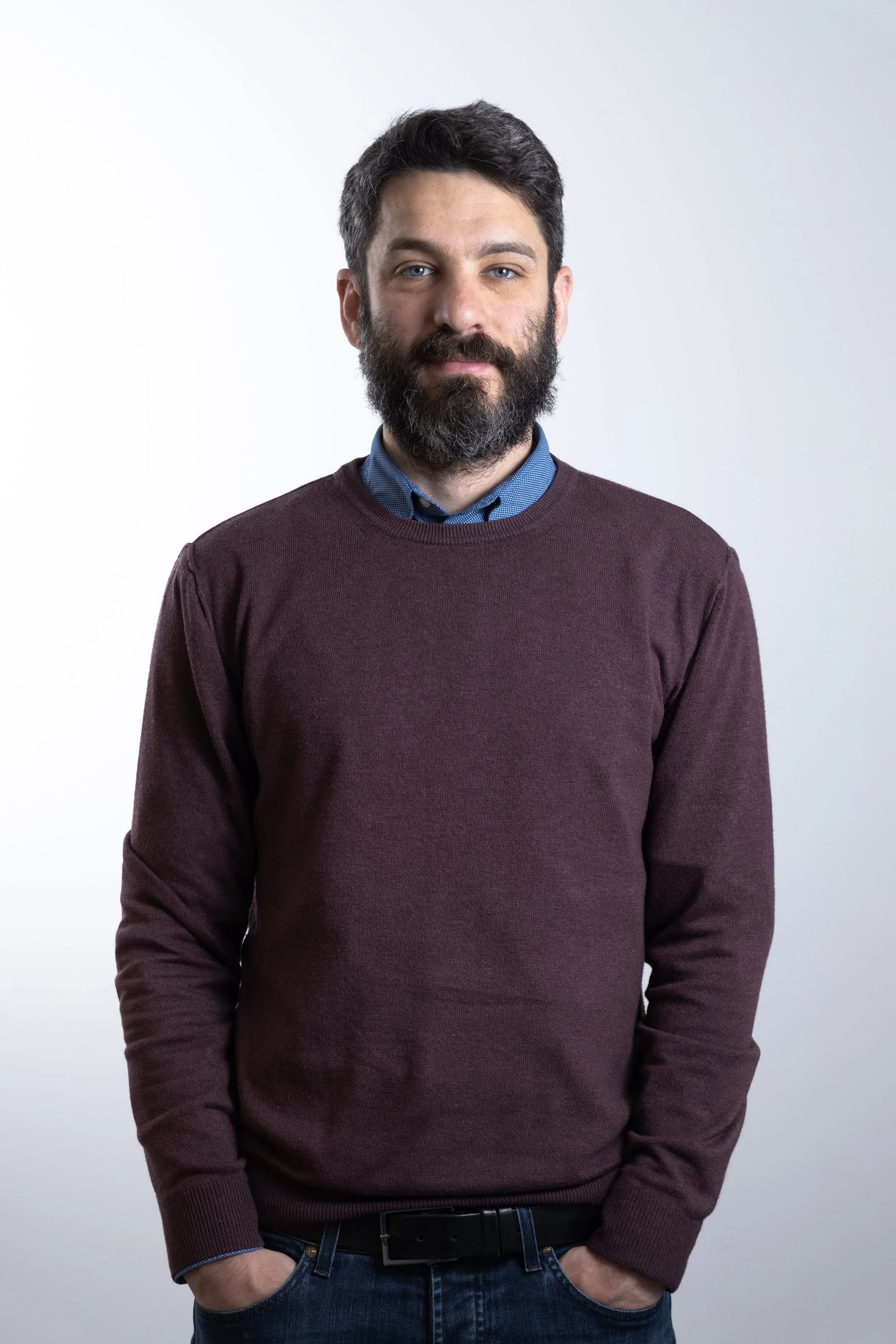 Uomo con barba, capelli castani, indossando una maglietta blu sotto un maglione marrone, con le mani nelle tasche.