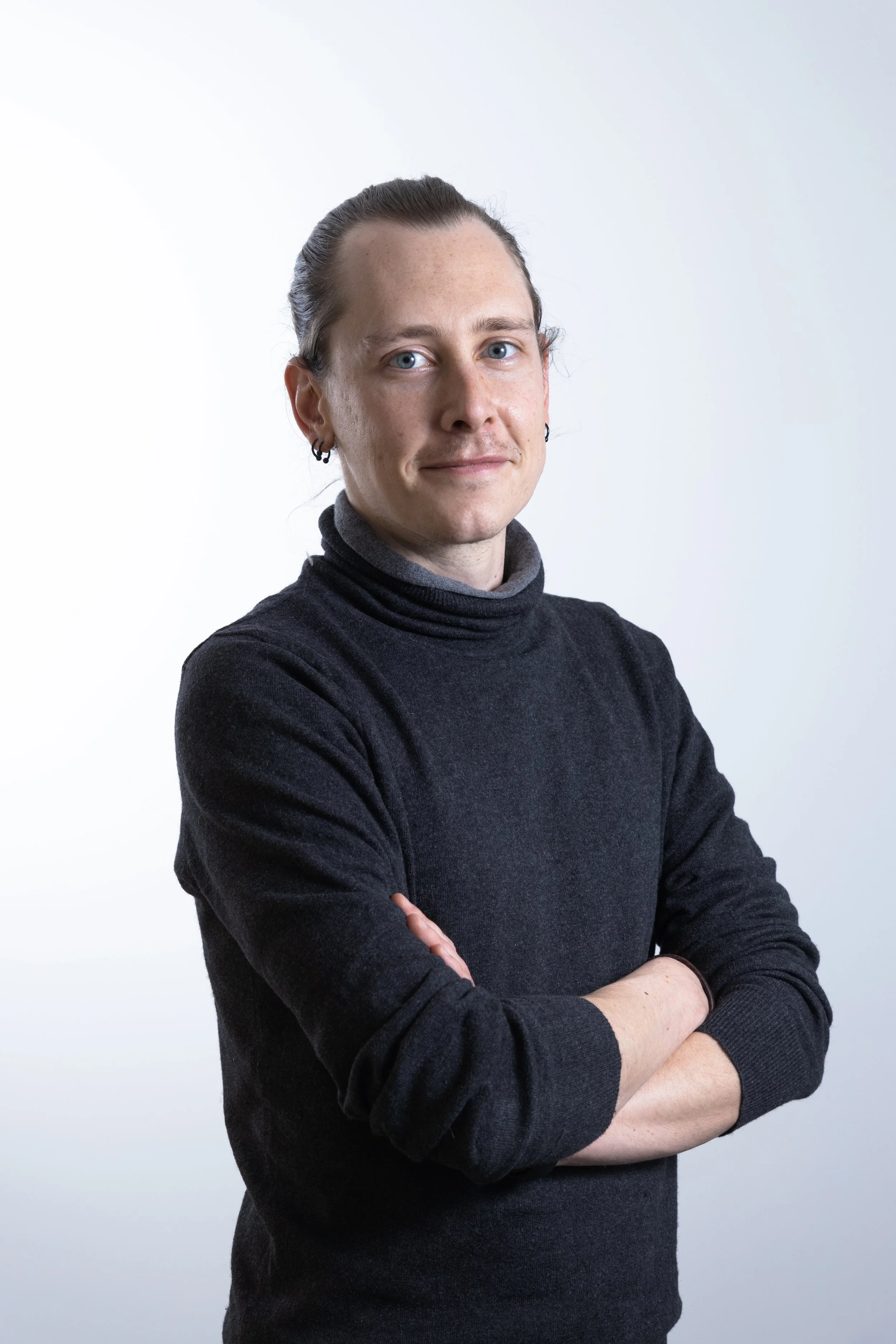 Ragazzo con capelli raccolti, indossa una maglia dolcevita nera, posa con le braccia incrociate davanti a uno sfondo bianco.