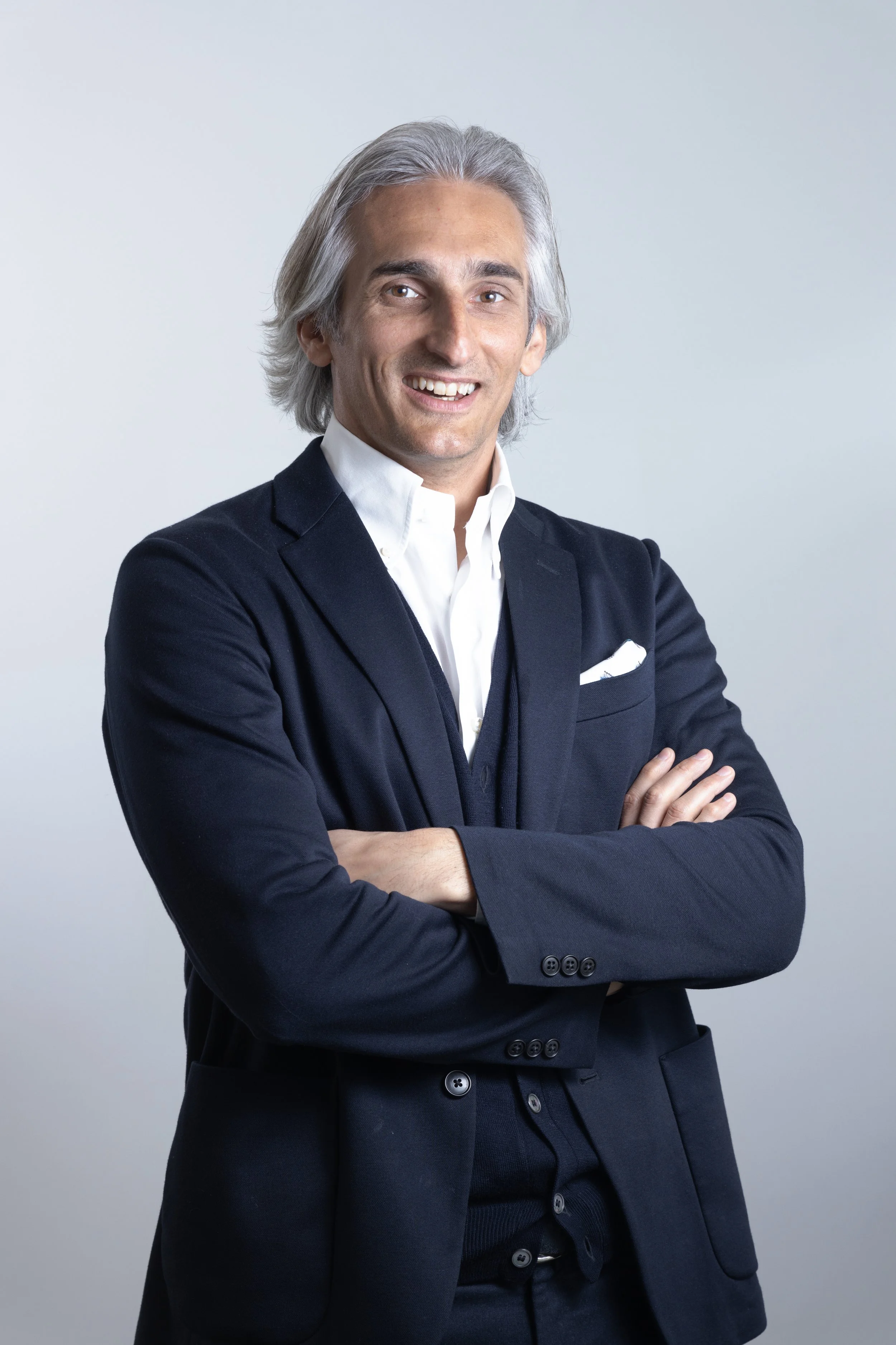 Uomo con capelli grigi, in abito blu scuro e camicia bianca, che sorride e ha le braccia incrociate.