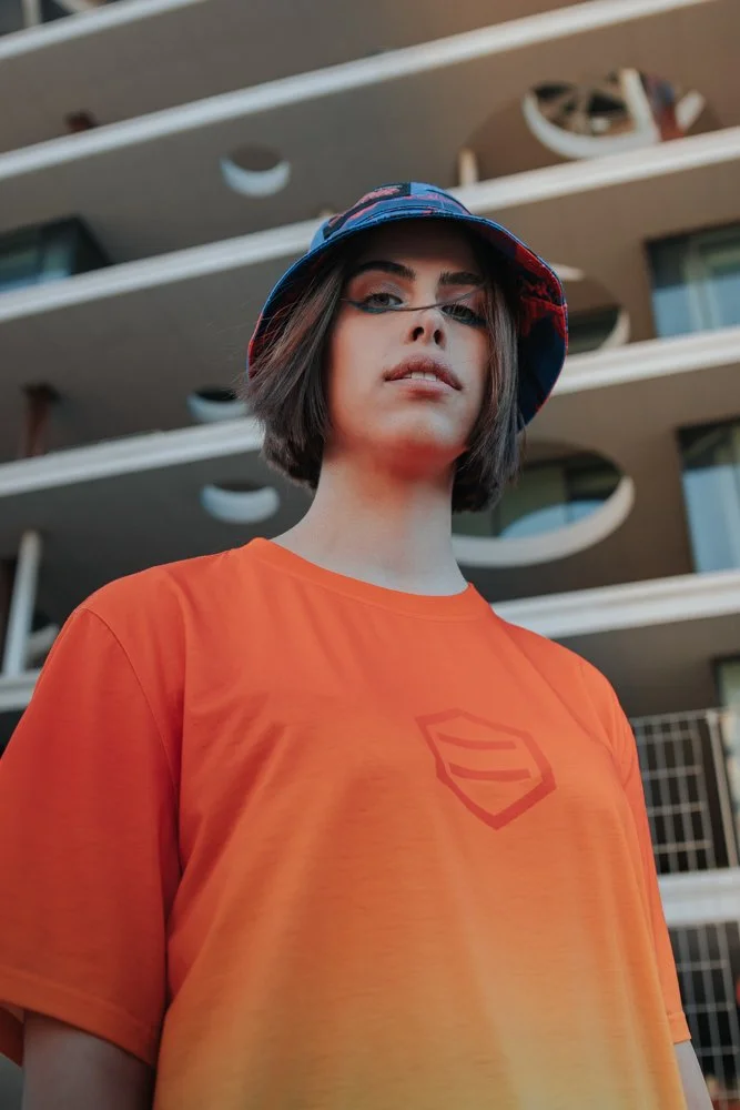 Ragazza con cappello colorato e t-shirt arancione con logo, in piedi davanti a un edificio moderno con balconi curvi.