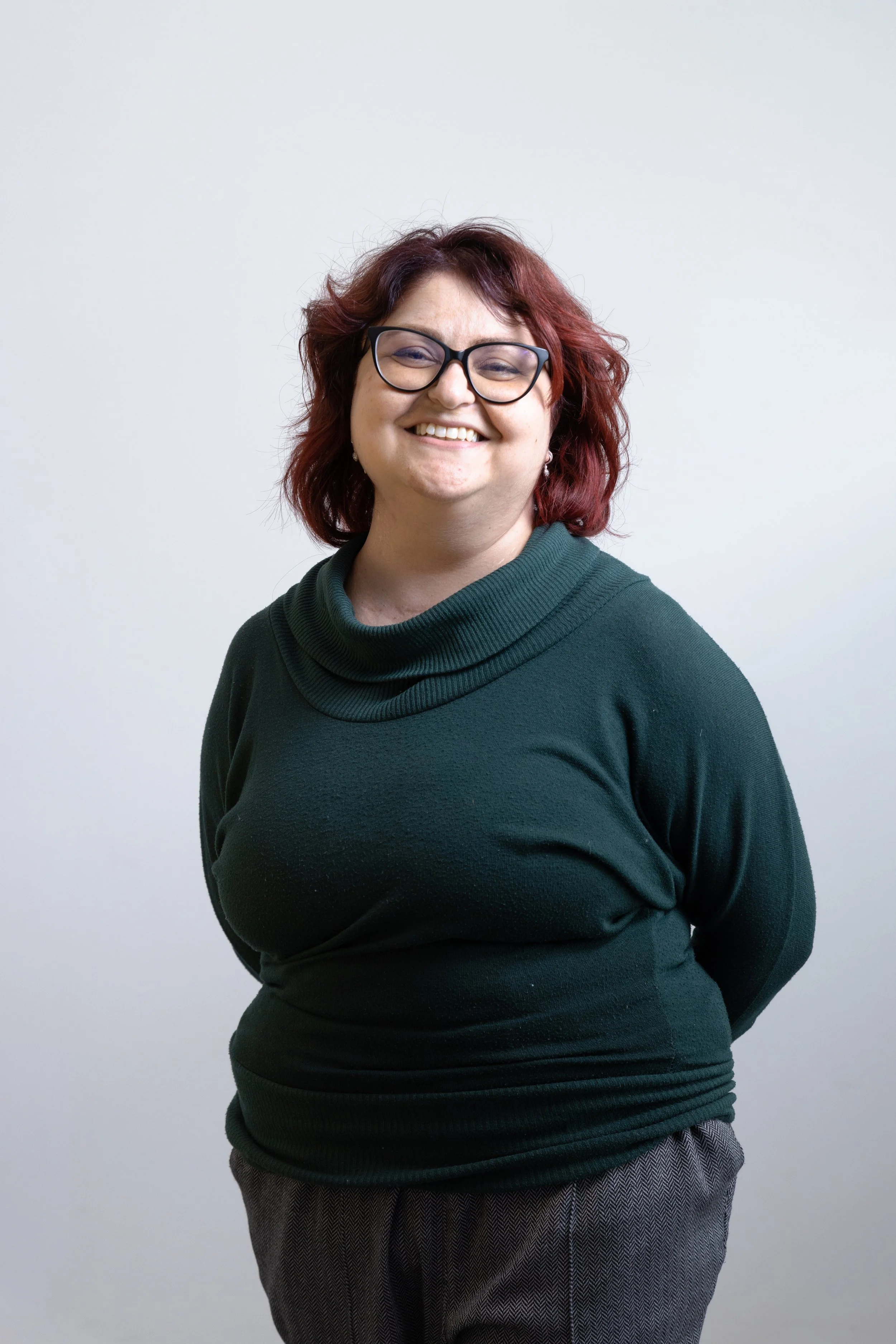 Donna sorridente con capelli ricci e occhiali, indossa una maglia verde scuro e pantaloni grigi, posa contro uno sfondo bianco.