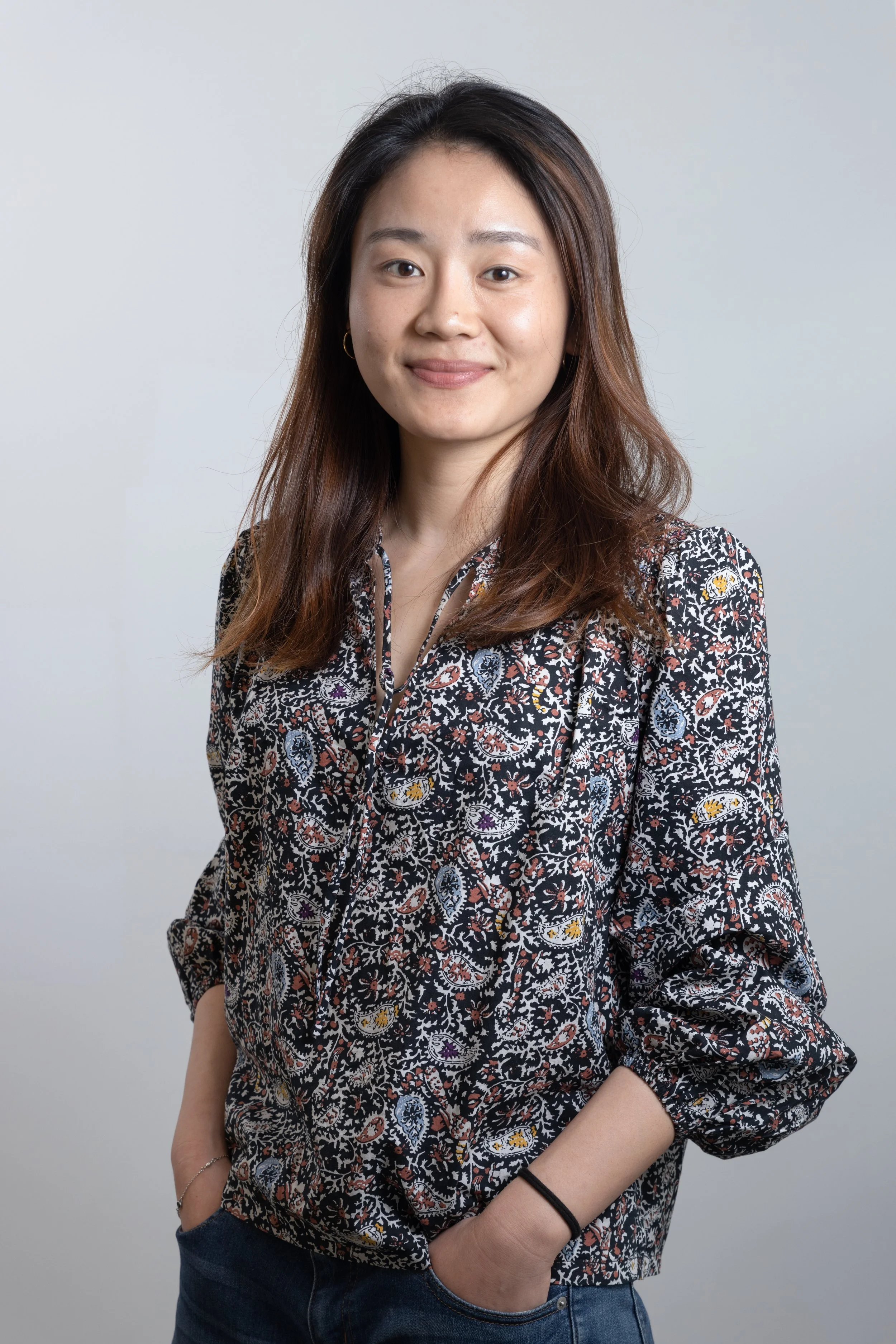 Ragazza sorridente con capelli castani, indossa una camicia a fantasia e jeans, porta le mani nelle tasche.