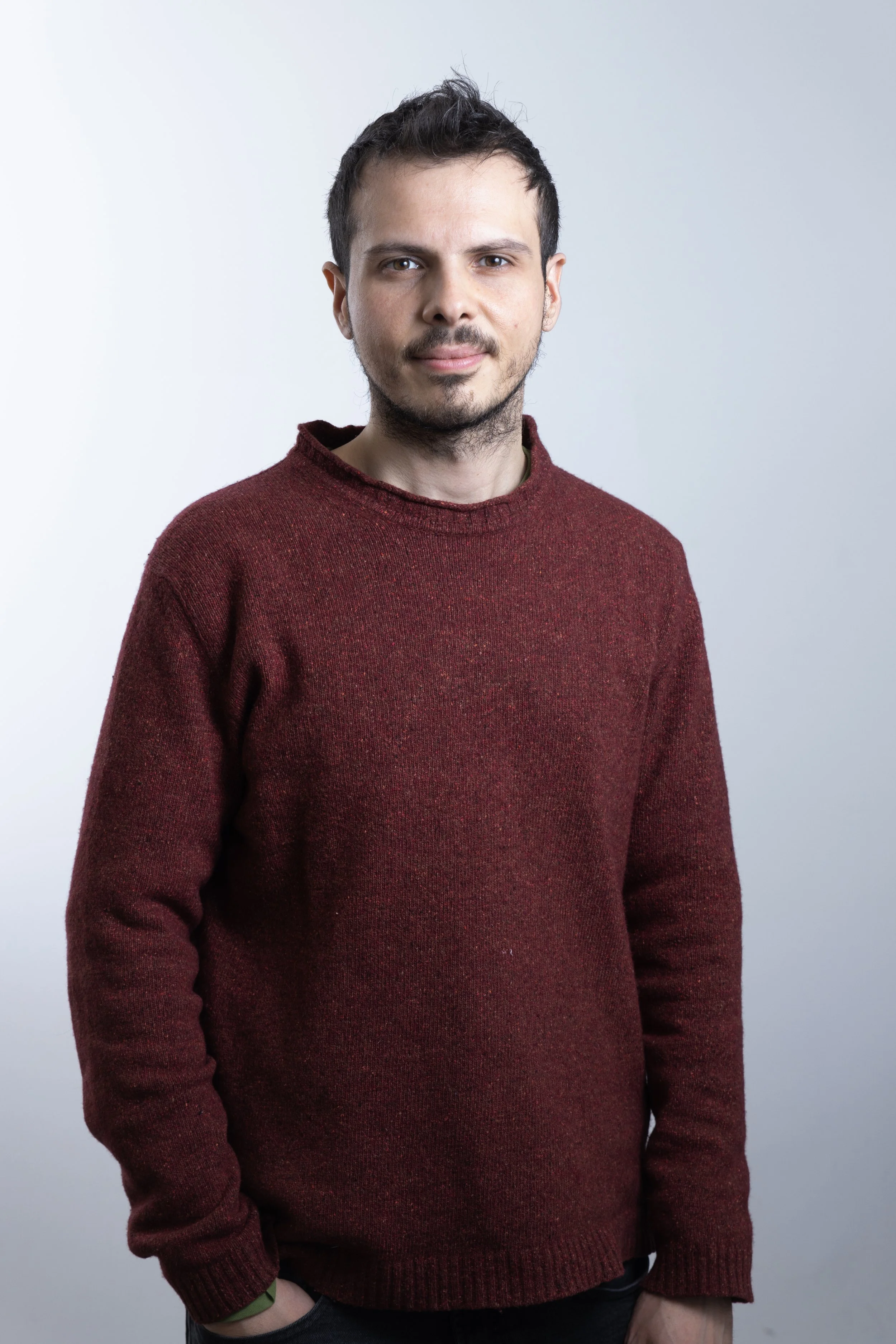 Un uomo giovane con capelli corti e barba, indossa un maglione rosso e guarda verso la fotocamera