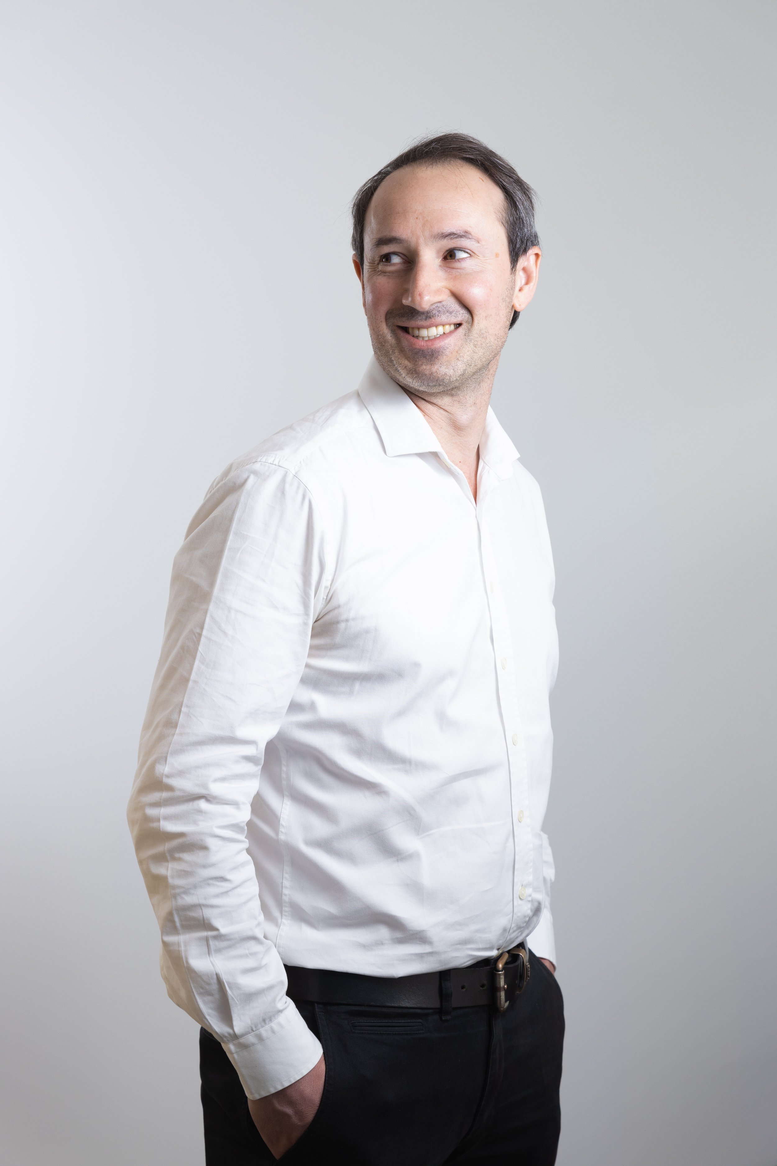 Uomo che indossa una camicia bianca e pantaloni neri, sorridente, con le mani in tasca, su sfondo bianco.