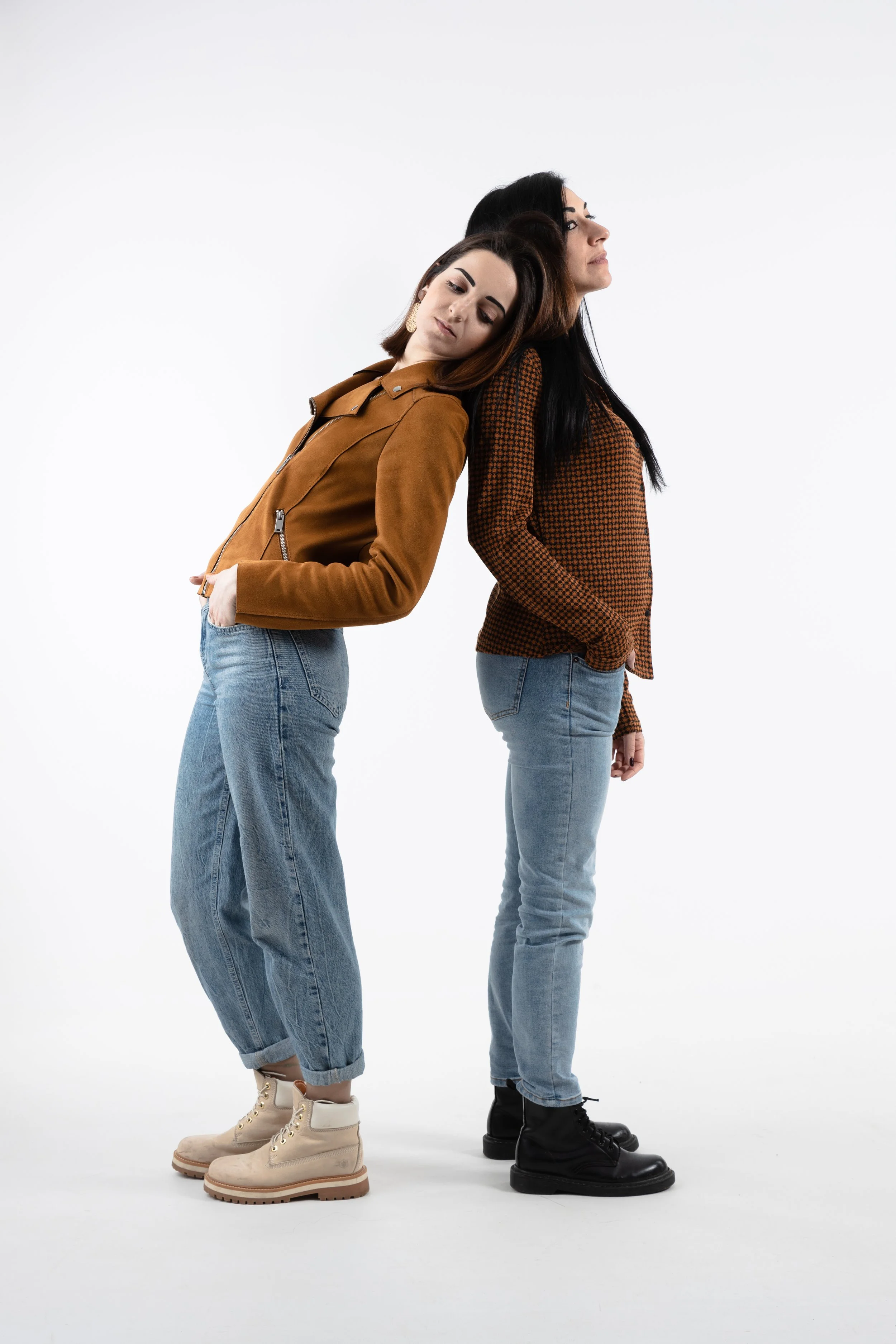 Due donne sono appoggiate le schiene l'una contro l'altra, con occhi chiusi. Una indossa una giacca marrone e jeans e l'altra una maglietta a quadri e jeans, entrambe con scarpe da stivali. La foto è scattata in uno studio con sfondo bianco.