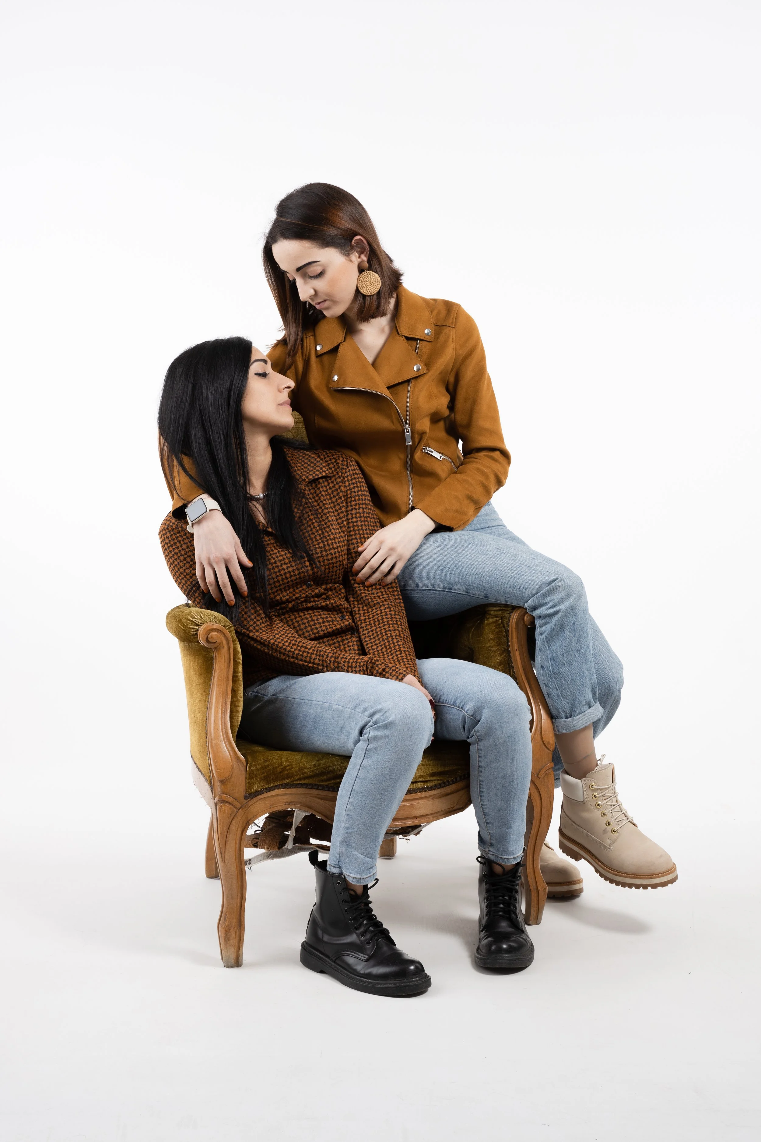 Due donne giovani, una seduta e l'altra in piedi, si abbracciano con un sorriso affiatato contro uno sfondo bianco.