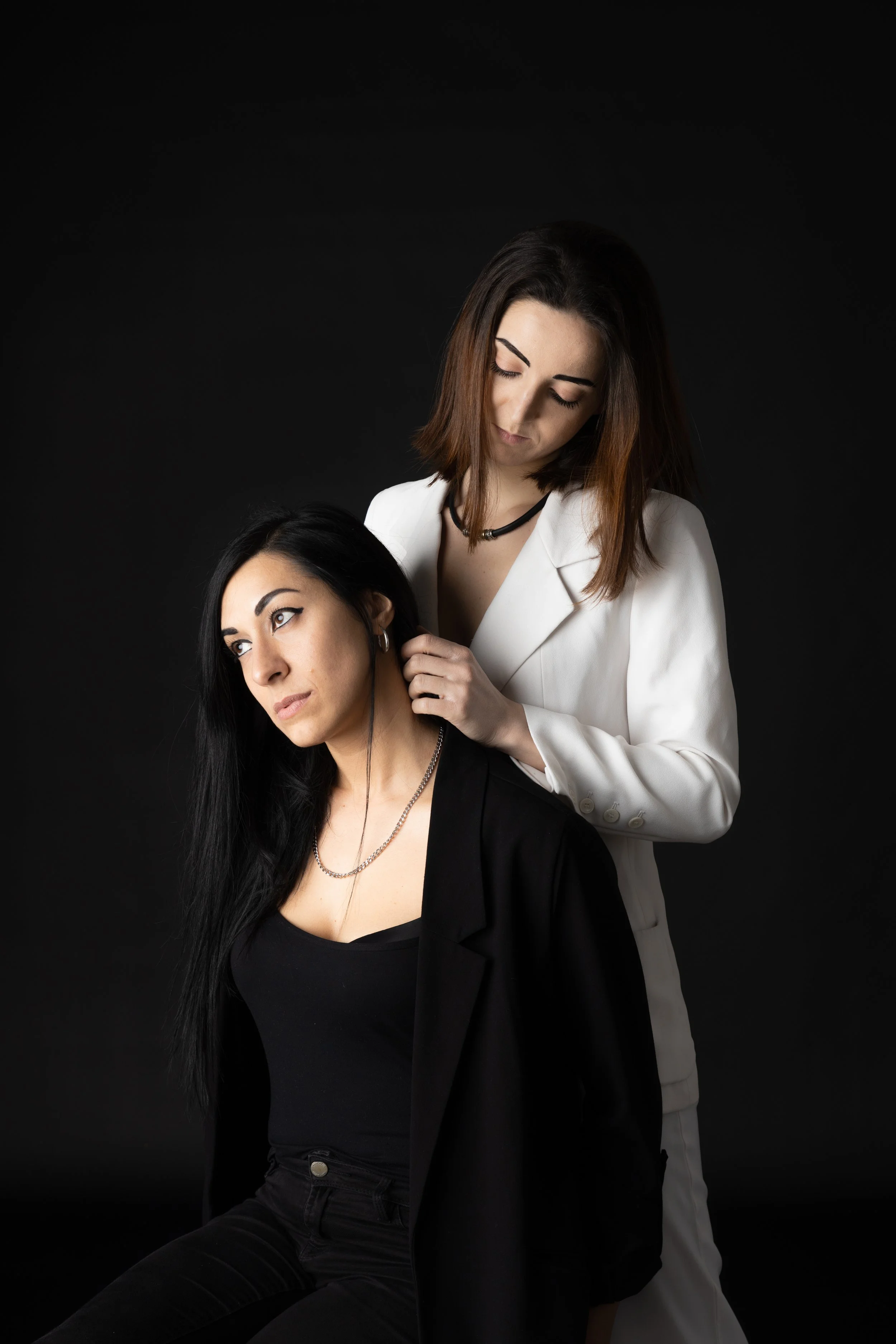 Due donne, una in abiti neri e l'altra in abiti bianchi, sedute e in posa contro uno sfondo scuro, con la donna in nero che indossa auricolari.