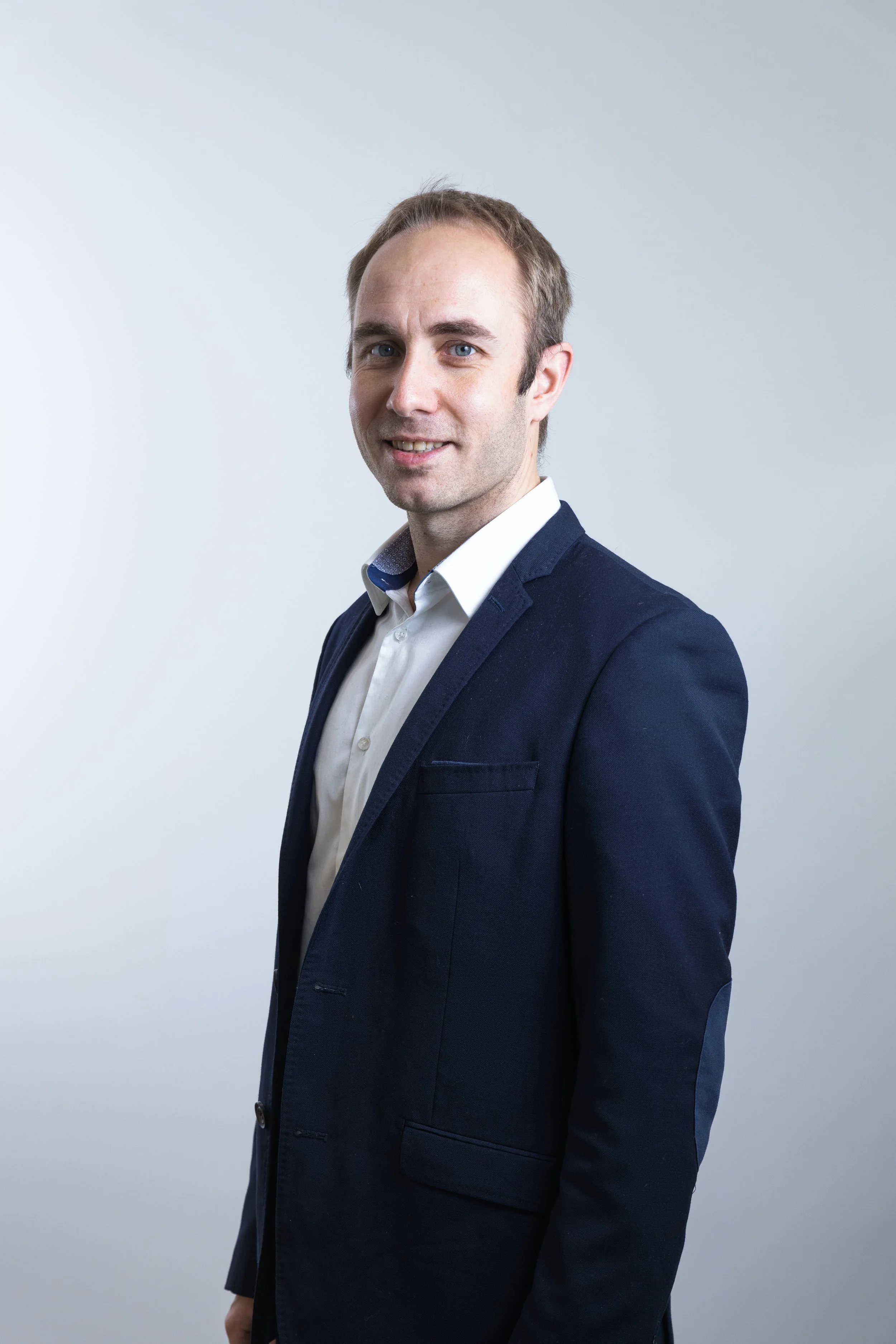 Uomo in abito blu scuro con camicia bianca, pose di profilo contro sfondo neutro.