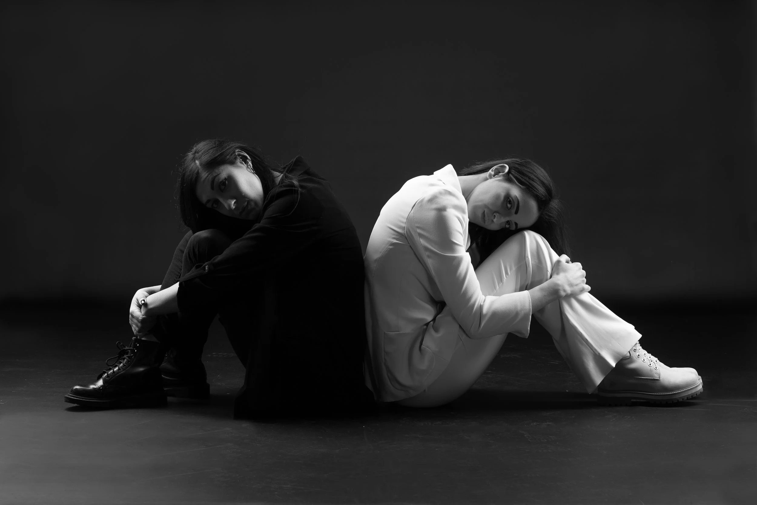 Due donne sedute a terra con le gambe piegate, una vestita di nero e l'altra di bianco, in un ambiente scuro, in bianco e nero.