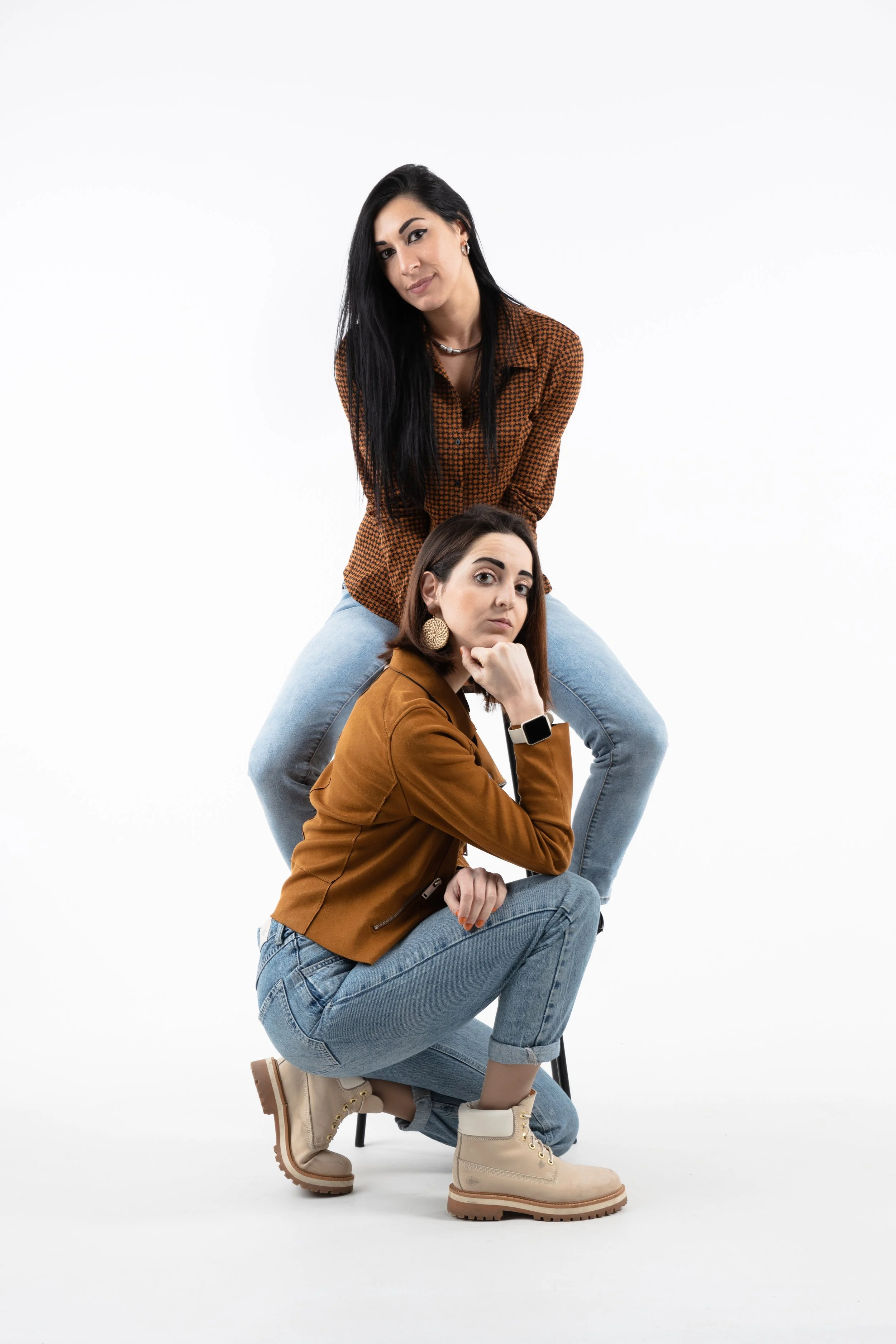 Due donne giovani si trovano una sopra l'altra in pose di pensiero, una indossa una giacca marrone e jeans, l'altra una camicia a quadri e jeans, entrambe con scarpe da trekking beige.