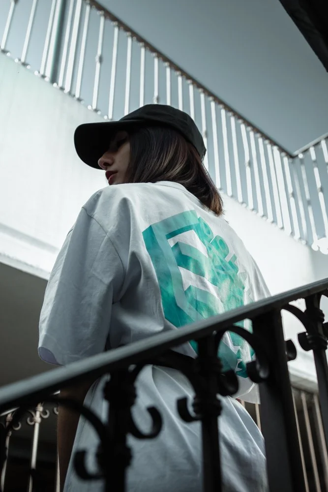 Donna con cappello nero indossa una maglietta bianca con disegno in tonalità di azzurro, vista dall'alto in un ambiente con ringhiera in ferro e luce naturale