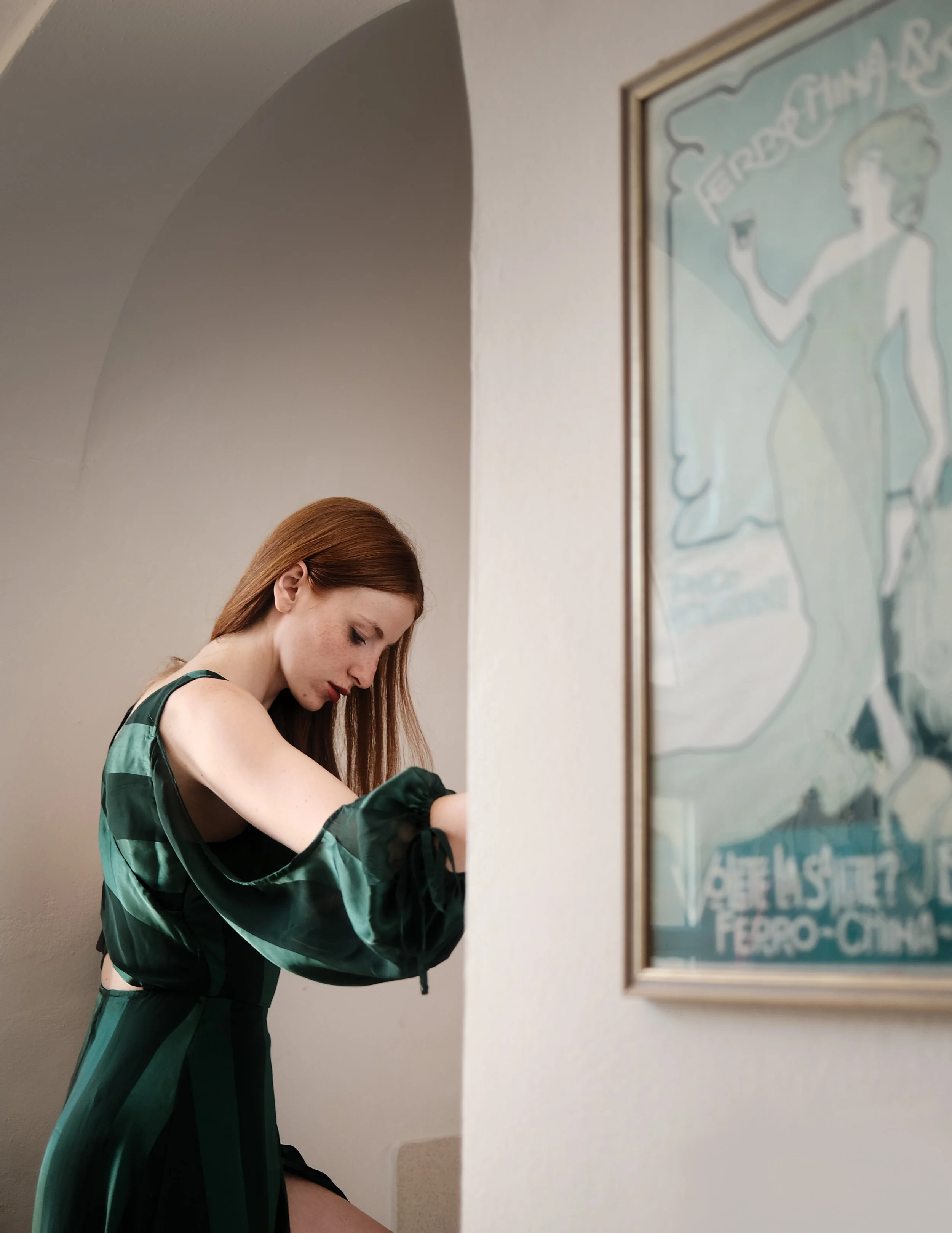 Una ragazza dai capelli castani con indumenti verdi seduta di profilo vicino a un muro, con un poster vintage sull'arte e la cultura. È concentrata e sembra che stia scrivendo o disegnando.