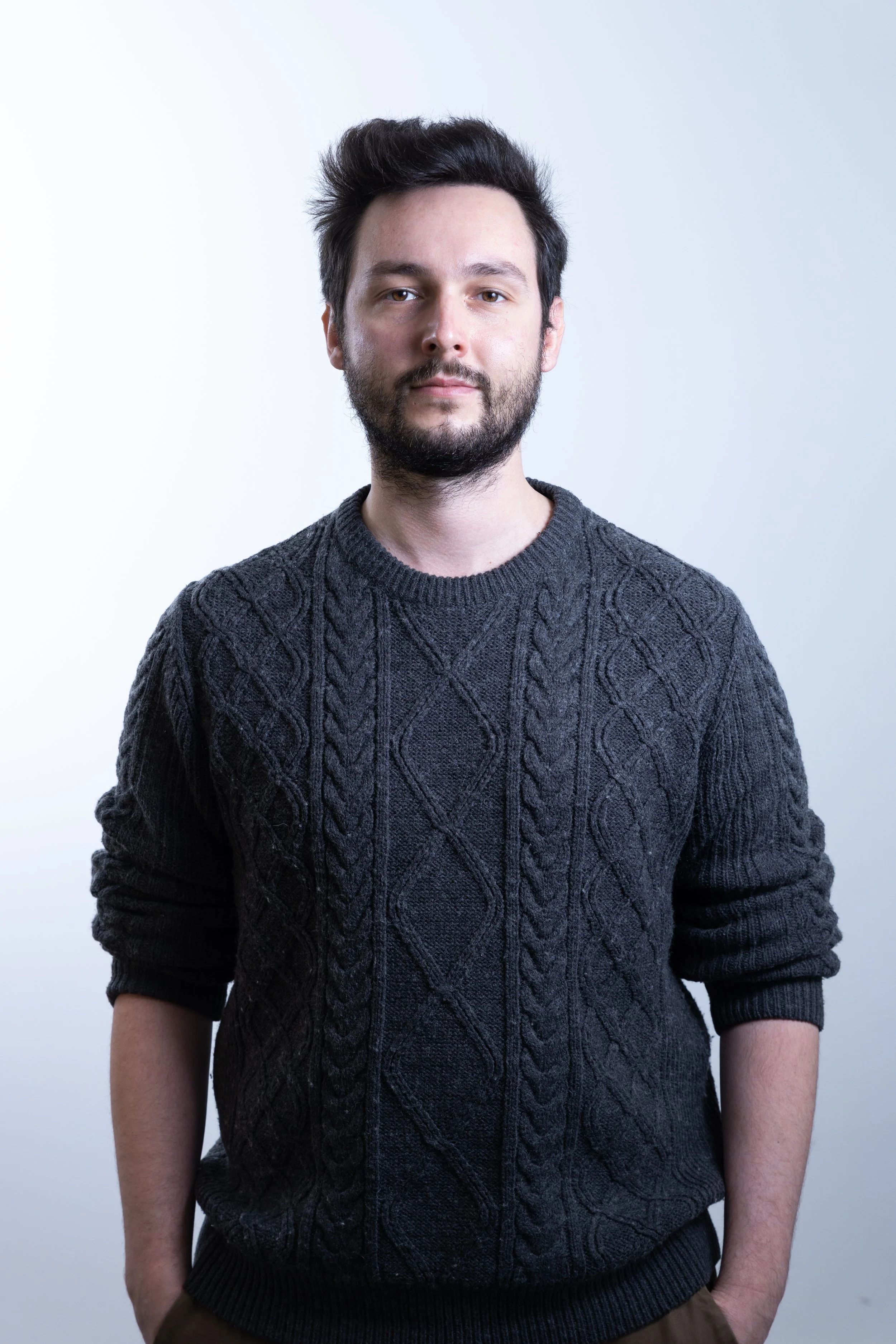 Ritratto di un uomo con capelli corti e barba, indossa un maglione grigio scuro con motivo a treccia, sfondo bianco