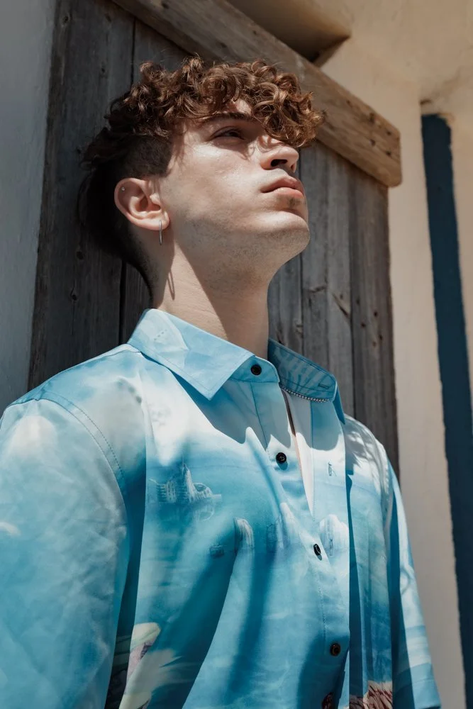Ragazzo con capelli ricci e abbigliamento con stampa di un paesaggio urbano, che si affaccia su un muro di legno, sotto una luce solare intensa.
