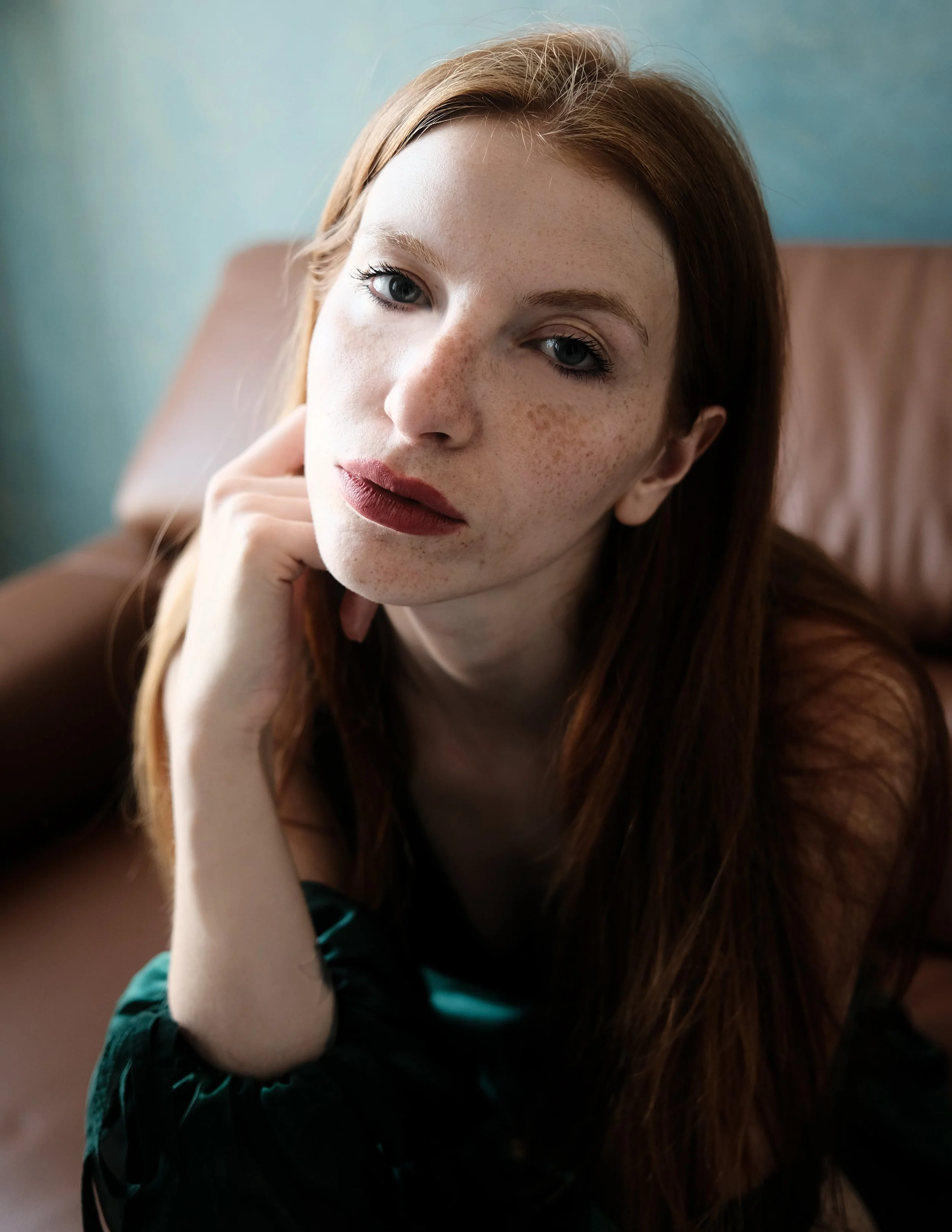 Ragazza con capelli rossi, viso con lentiggini, riposa il mento sulla mano e guarda verso la fotocamera in un ambiente interno.