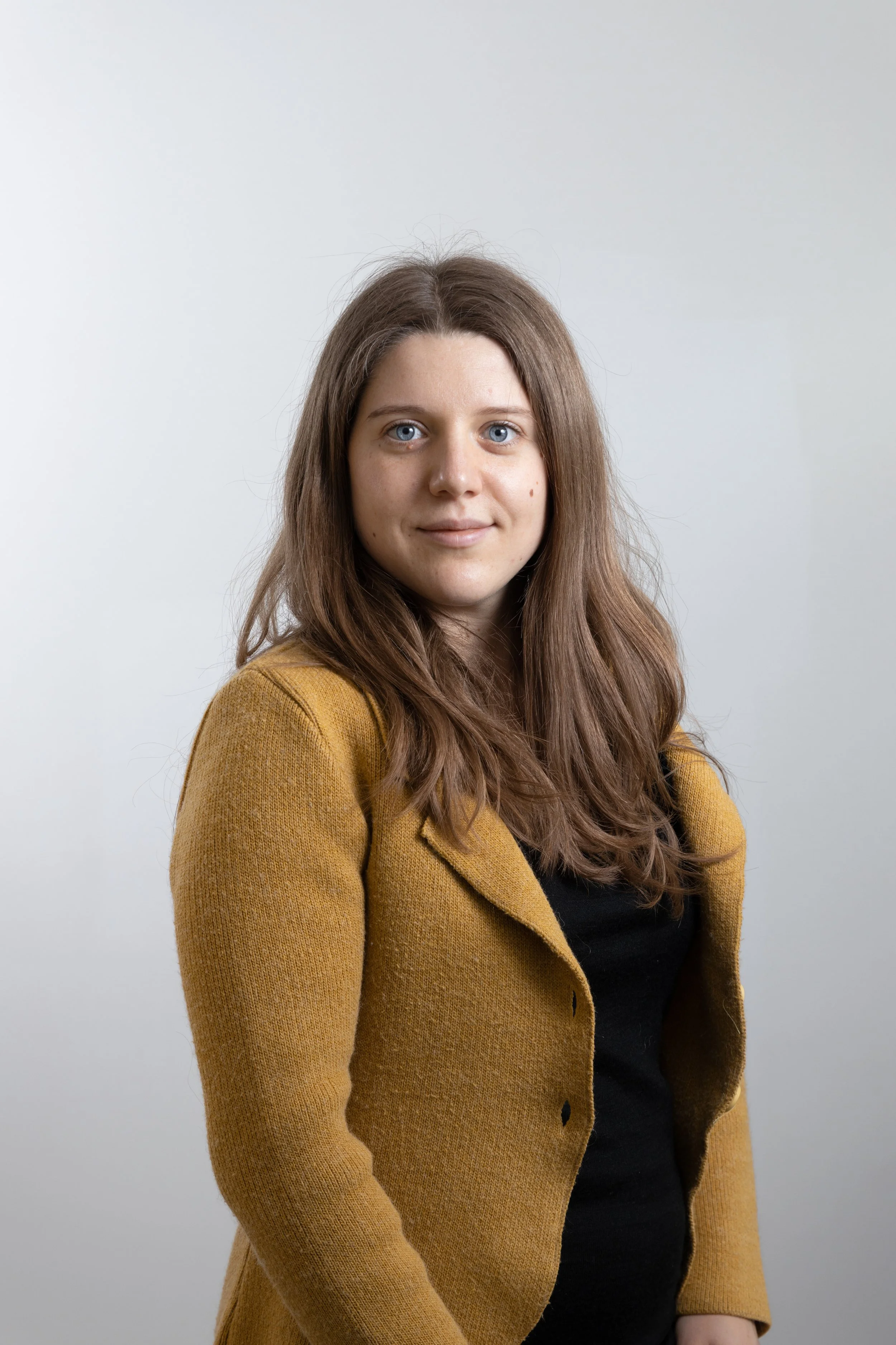 Una giovane donna dai capelli castani e occhi azzurri, indossa una giacca marrone e una maglietta nera, posa di fronte a uno sfondo bianco.