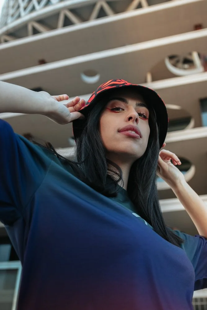 Una donna con cappello a tesa larga nera e rossa, capello nero e capelli lunghi, indossa una maglietta blu scuro, si trova davanti a un edificio moderno con balconi curvi