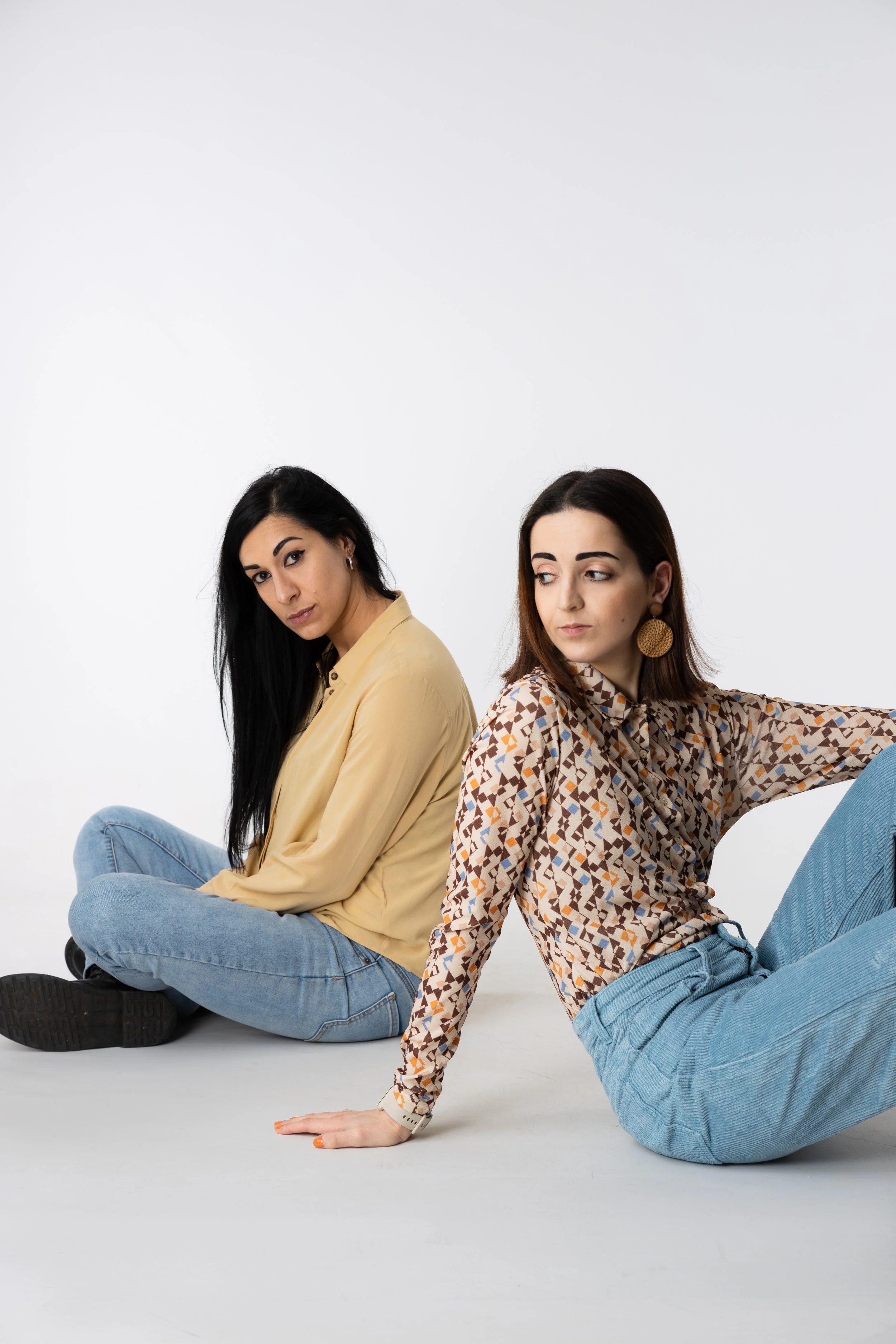 Due donne sedute a terra contro uno sfondo bianco, una con camicia beige e jeans, l'altra con camicia stampata e pantaloni azzurri.