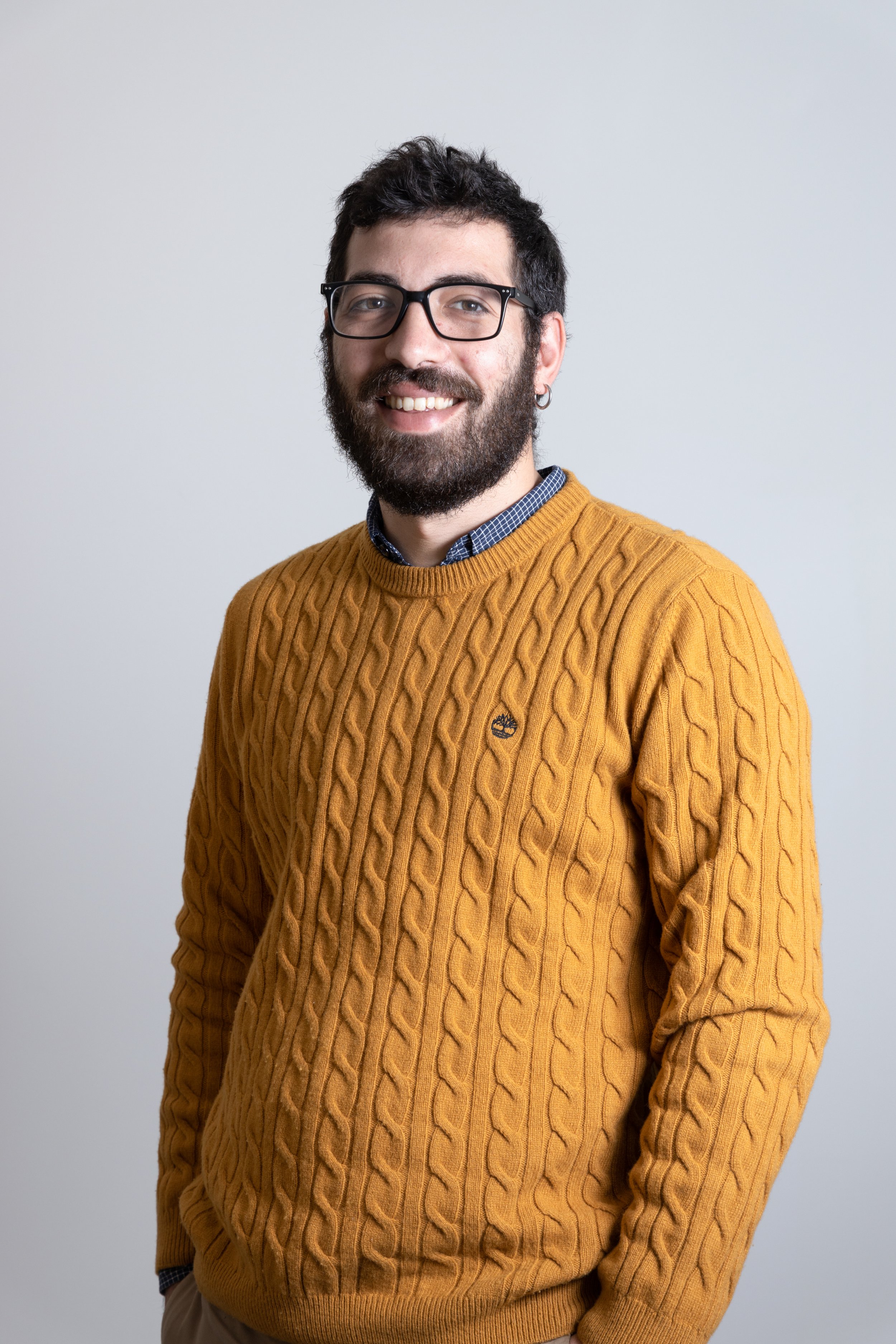 Uomo sorridente con barba, occhiali e orecchino indossa un maglione giallo con motivo a treccia, sfondo bianco.