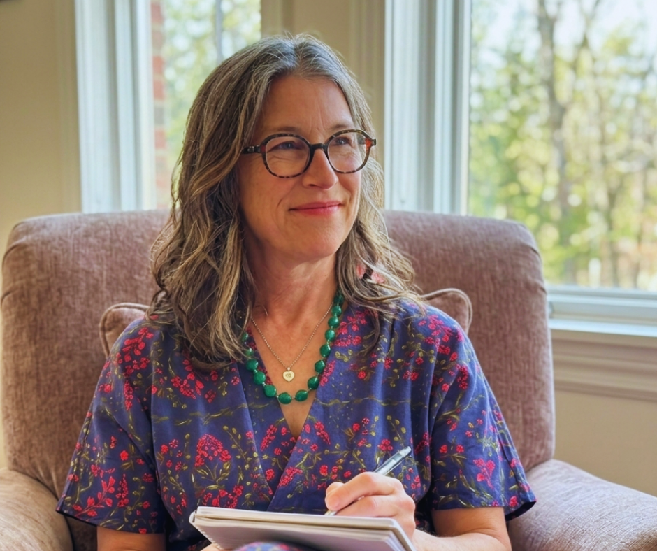 Heather Ramsey with shoulder-length gray hair, wearing glasses, a colorful floral dress, a green beaded necklace, and a small heart pendant, sitting in a beige armchair near a window with trees outside, holding a notebook and pen, smiling gently.