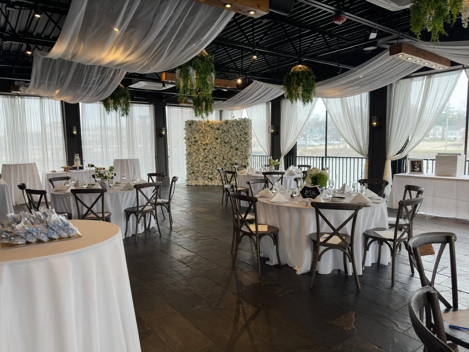 Wedding reception venue with round tables, white linens, floral wall, hanging greenery, and draped sheer curtains.