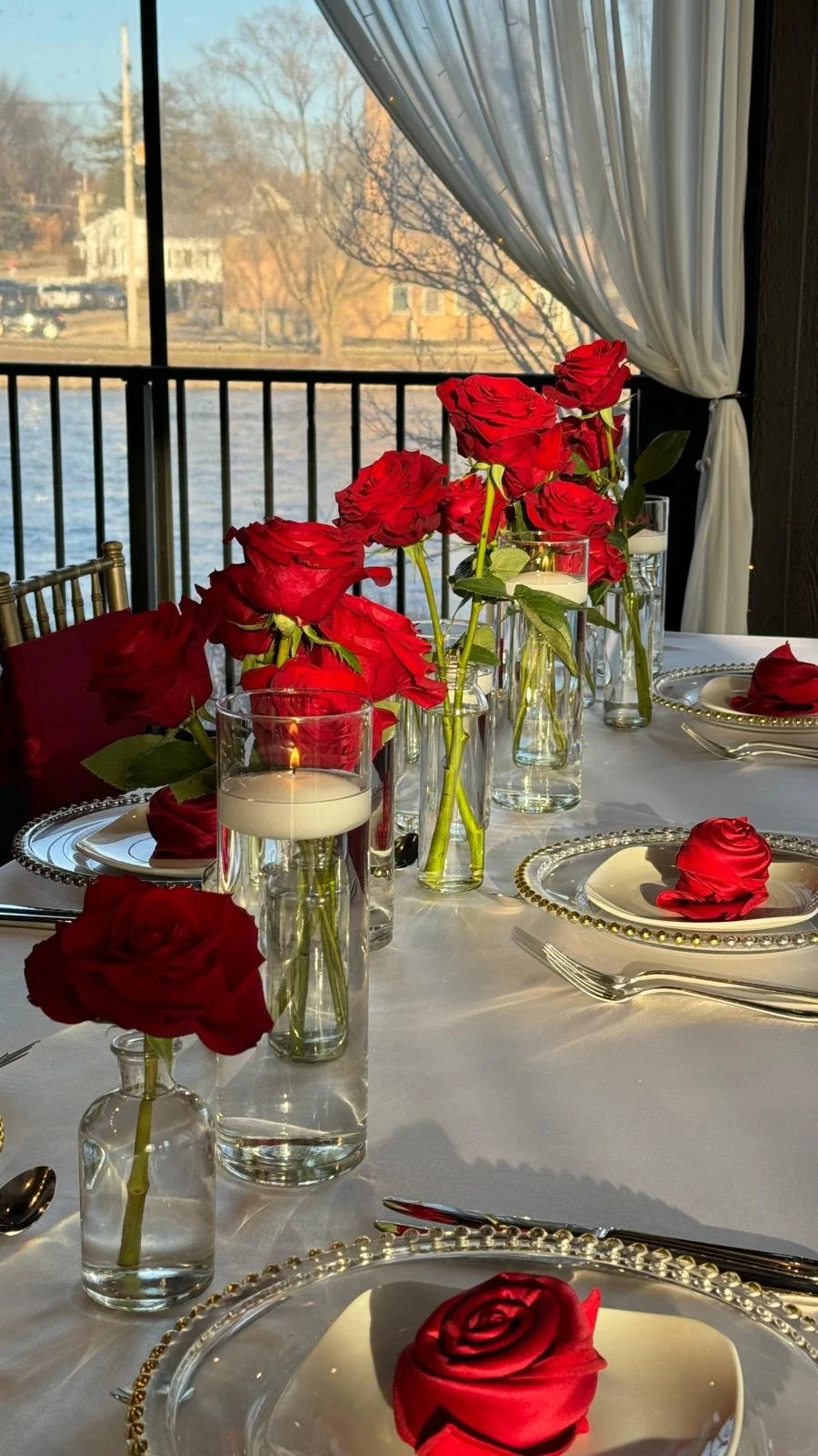 Elegant table setting with red roses, floating candles, white plates, and gold-rimmed chargers on a white tablecloth, near a window with sheer curtains.