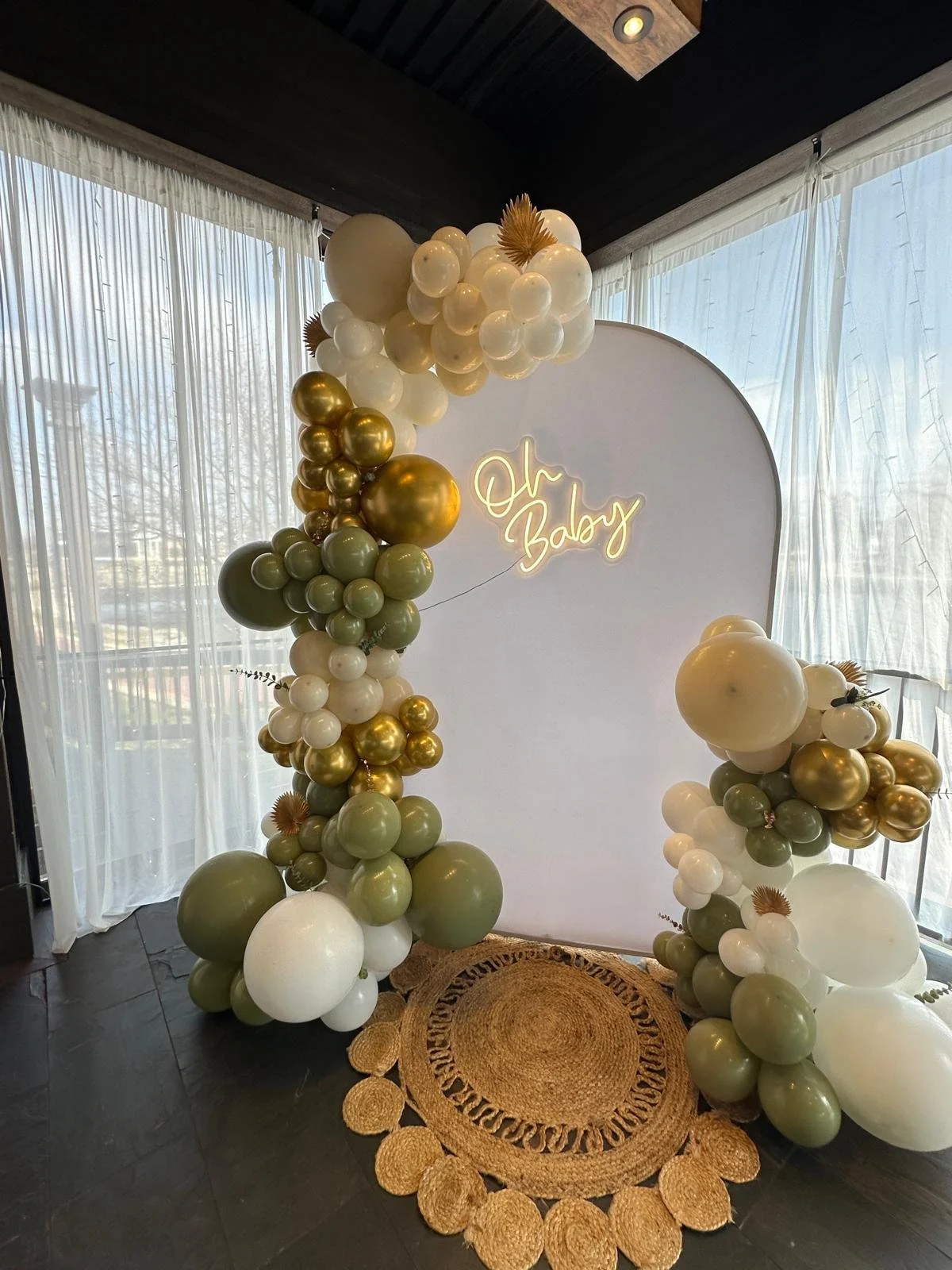 Baby shower setup with balloon arch, green and white balloons, "Oh Baby" neon sign, and round woven rugs.