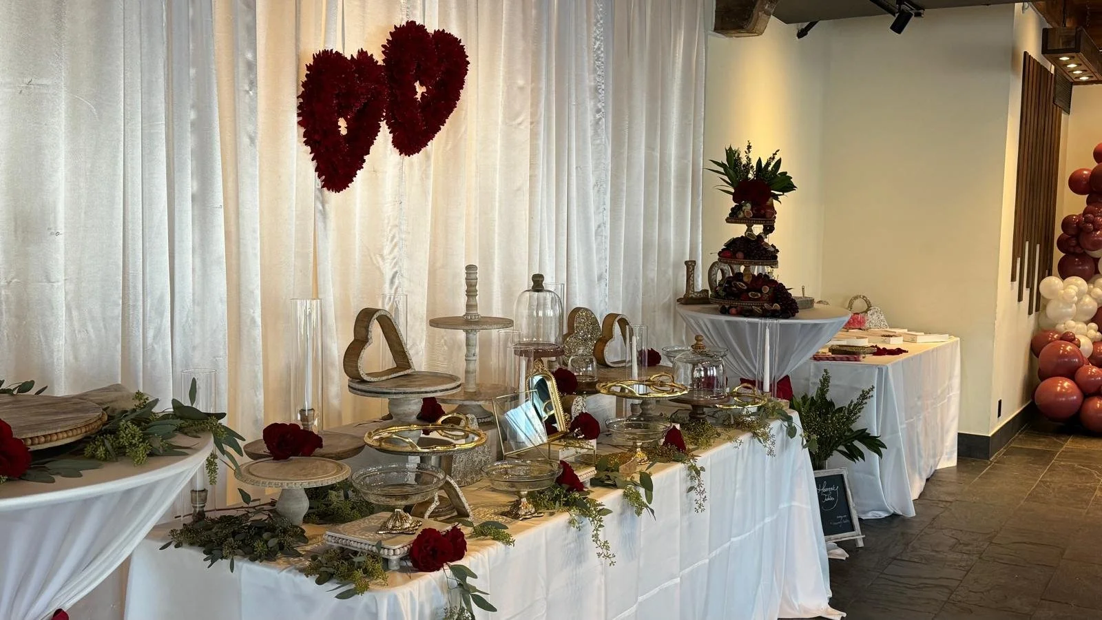 Wedding dessert table with wooden stands, glass cloches, red roses, greenery, wine-colored balloon arrangement, and heart-shaped floral decor.
