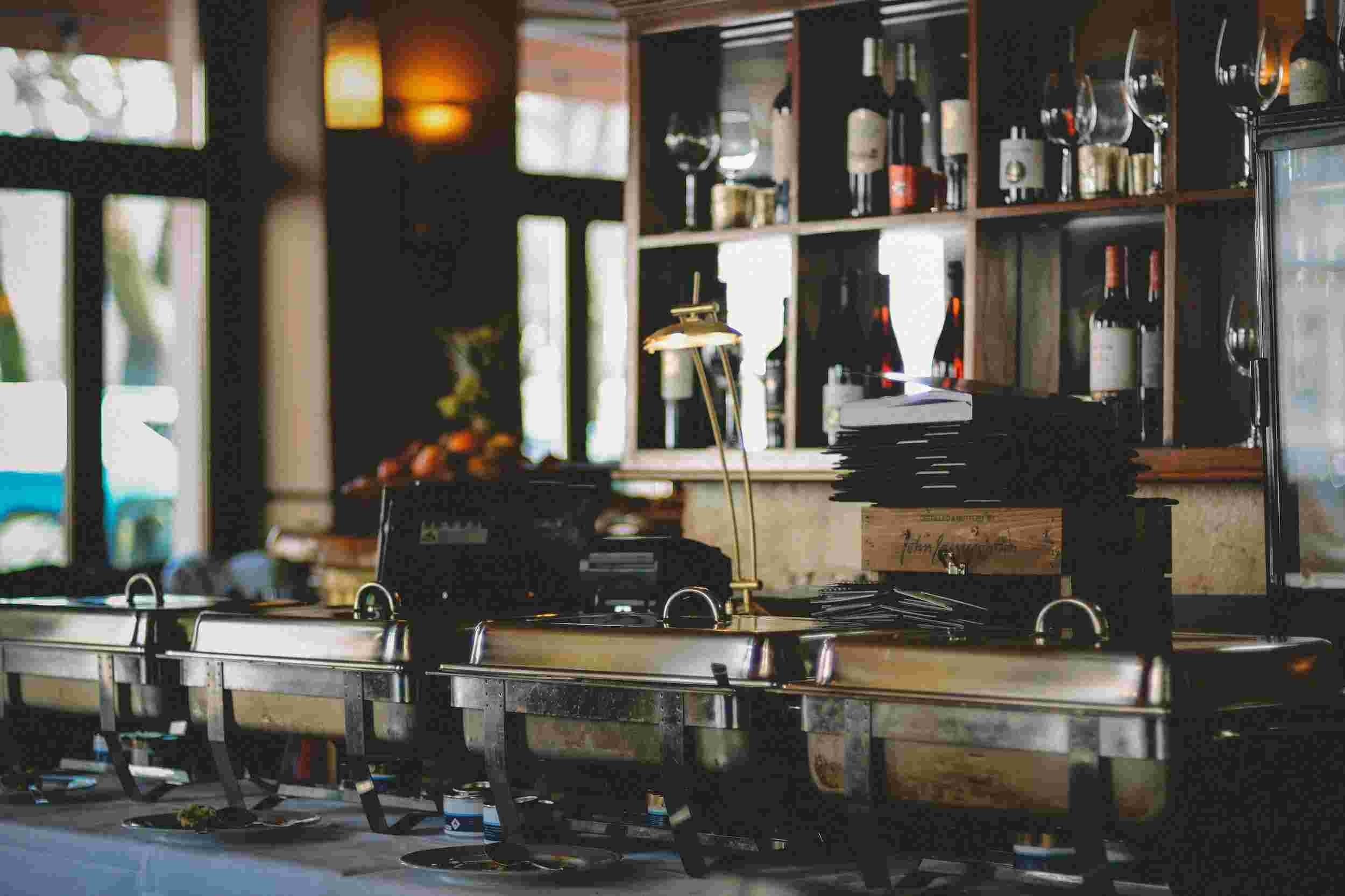 Restaurant buffet setup with chafing dishes, plates, and a shelf displaying wine bottles and glasses in the background.