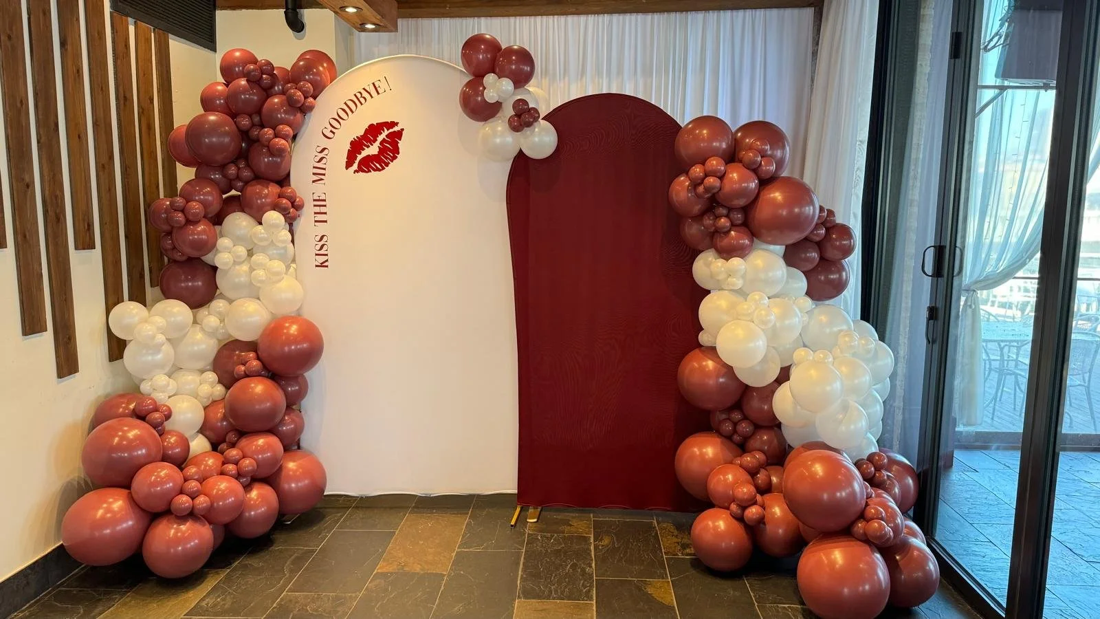 Balloon arch with maroon and white balloons, and a white panel with 'Kiss the Miss Goodbye!' and red lips graphic, for a bridal shower decoration.