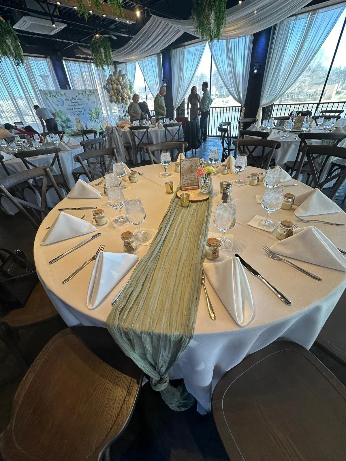 Elegantly set banquet table with white tablecloth, green table runner, and neatly arranged silverware. Surrounding tables and chairs are visible in a decorated venue with draped curtains and greenery. Guests are seen in the background near a balloon