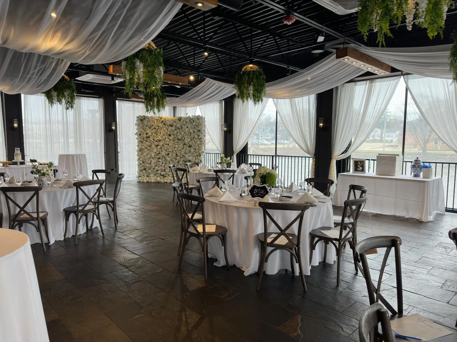 Elegant wedding reception venue with tables set for guests, white tablecloths, wooden chairs, ceiling drapery, green foliage, and a floral wall backdrop.