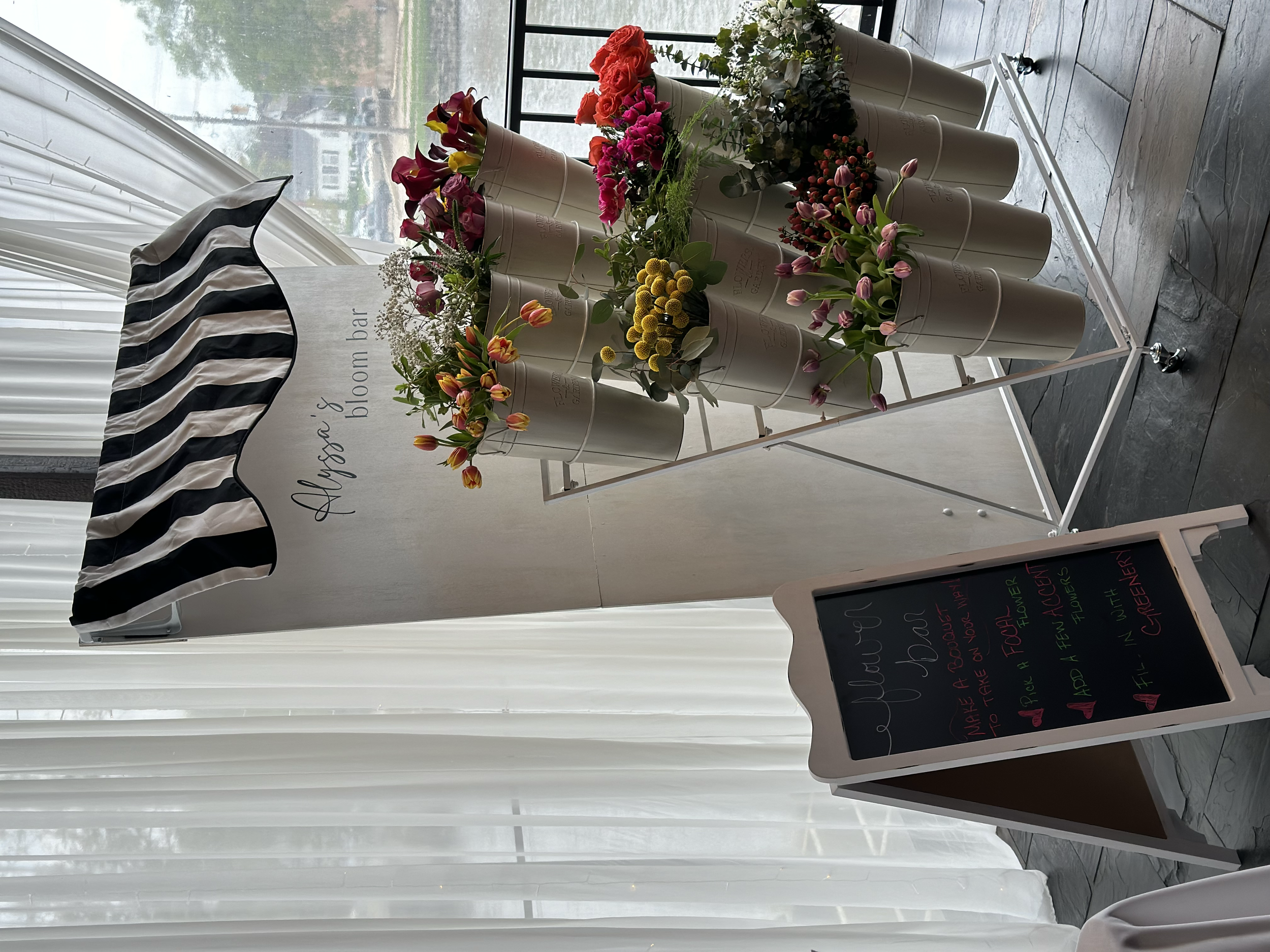 Flower bar setup with arranged floral bouquets in white buckets, featuring a black and white striped canopy and a chalkboard sign offering floral arrangements. Background includes sheer white curtains and glass window.