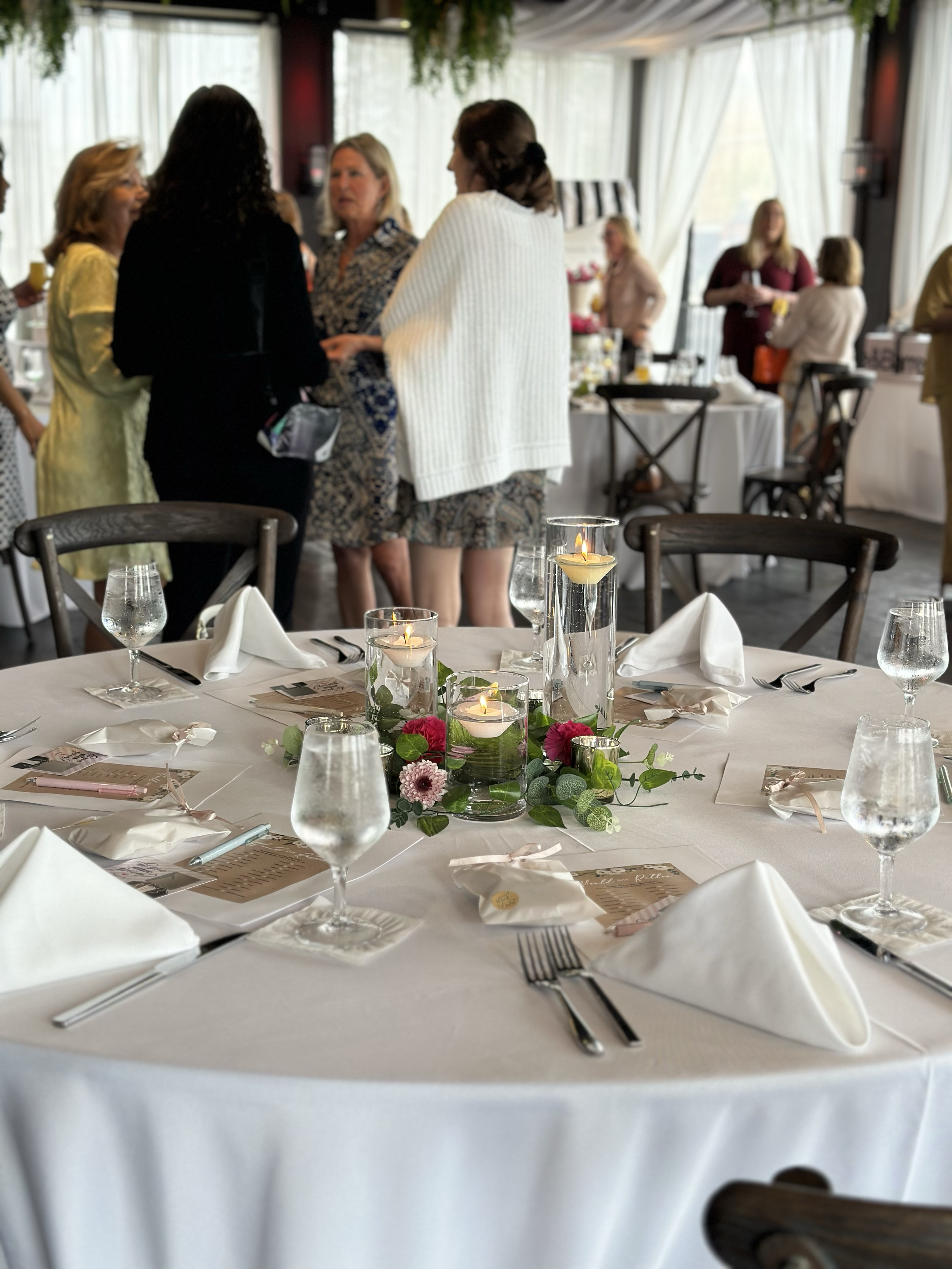 Group of people mingling at a formal event with a decorated round table in the foreground, featuring white tablecloth, floral centerpiece with candles, and neatly arranged water glasses and utensils.