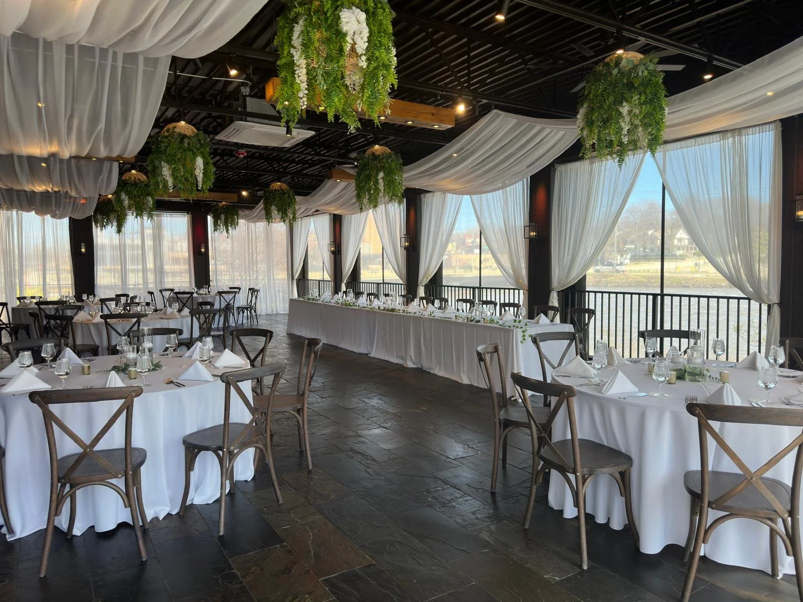 Elegant indoor wedding reception setup with round and long tables covered in white tablecloths, white draped ceilings, and hanging greenery. Large windows with sheer curtains overlook a scenic view.
