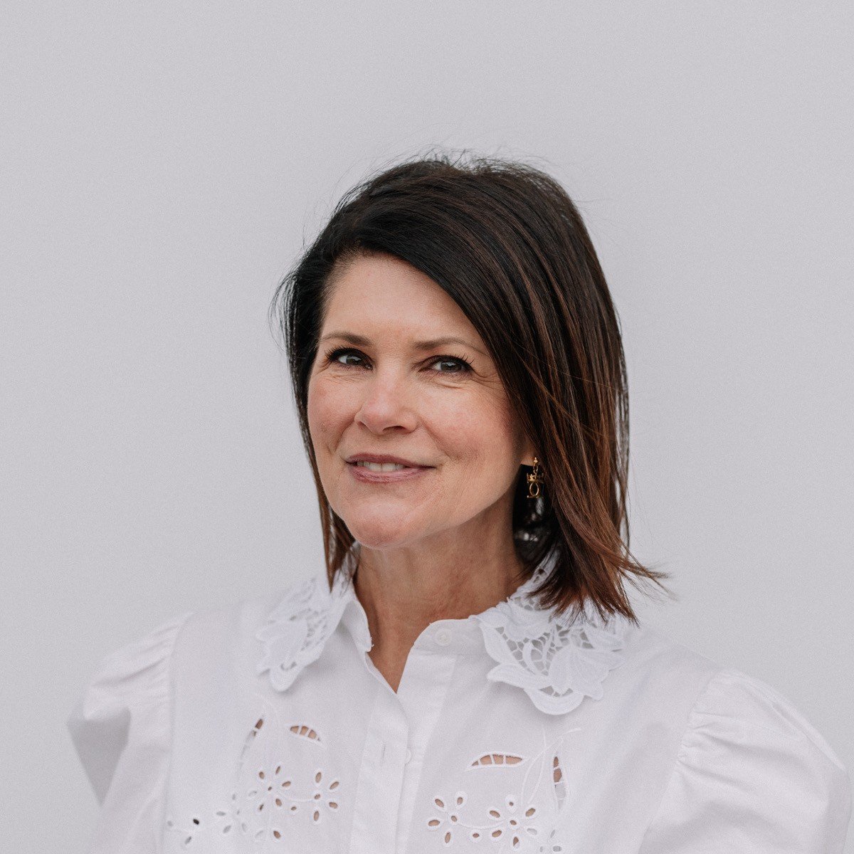 A woman with shoulder-length dark hair wearing a white embroidered blouse, smiling slightly against a plain gray background.