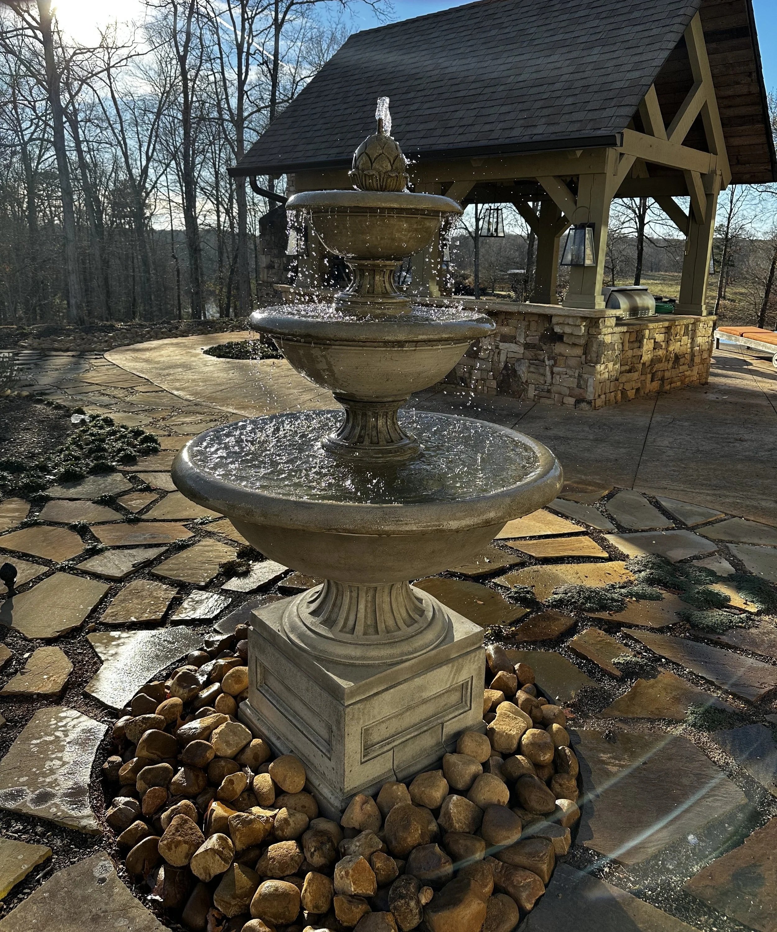 Three-tier stone water fountain on a flagstone patio near a covered outdoor kitchen pavilion.
