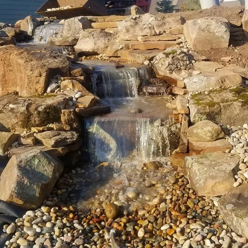 Close-up of a tiered stone waterfall cascading over natural rock ledges into a pebble-lined stream.