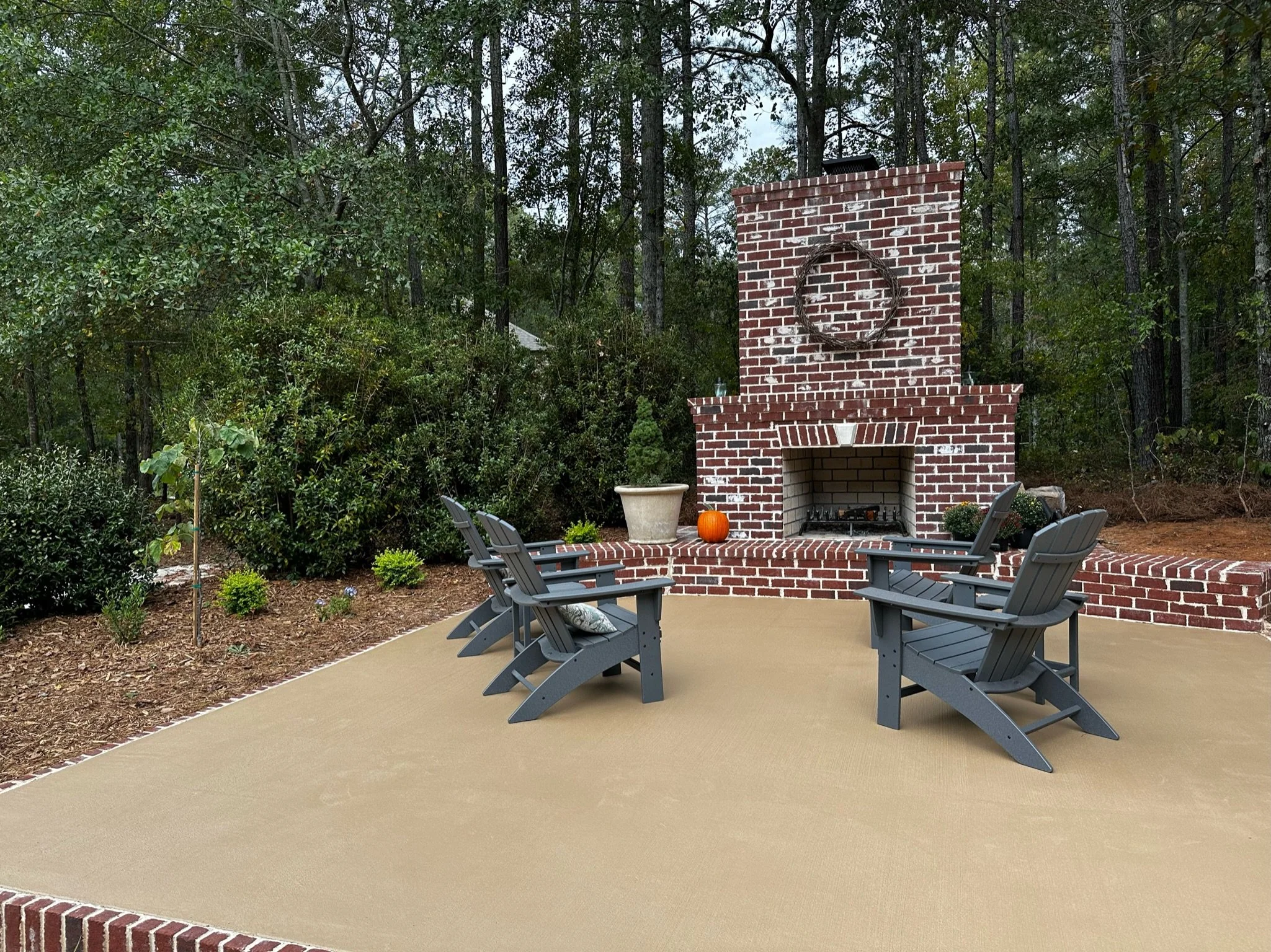 Concrete patio with brick outdoor fireplace and Adirondack chairs surrounded by trees.