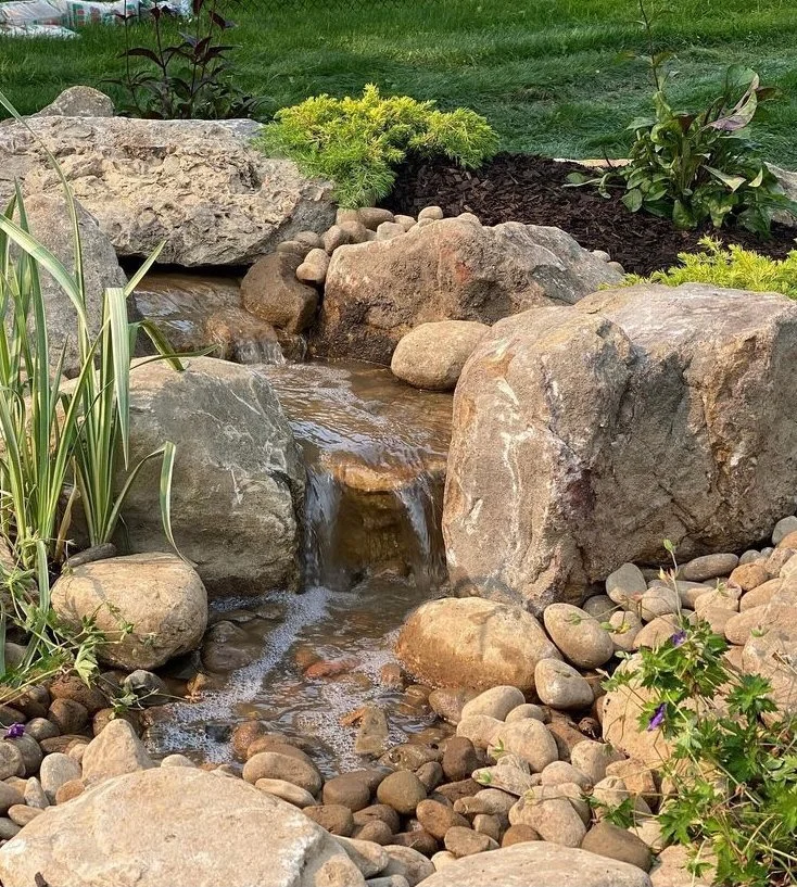 Boulder waterfall feature with cascading stream flowing through river rock and native grasses.