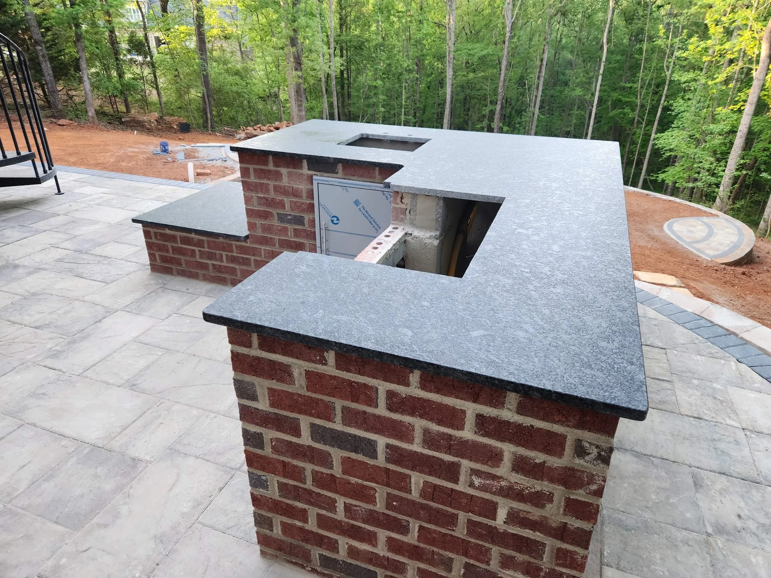 L-shaped brick outdoor kitchen with granite countertops and stainless steel appliance cutouts surrounded by wooded landscape.