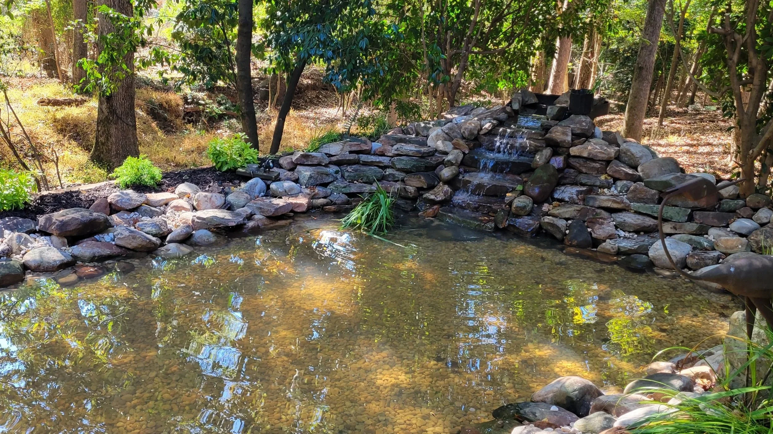 Natural stone waterfall flowing into a backyard pond with river rock edging and aquatic plantings by Southern Grounds Landscaping in Athens Georgia
