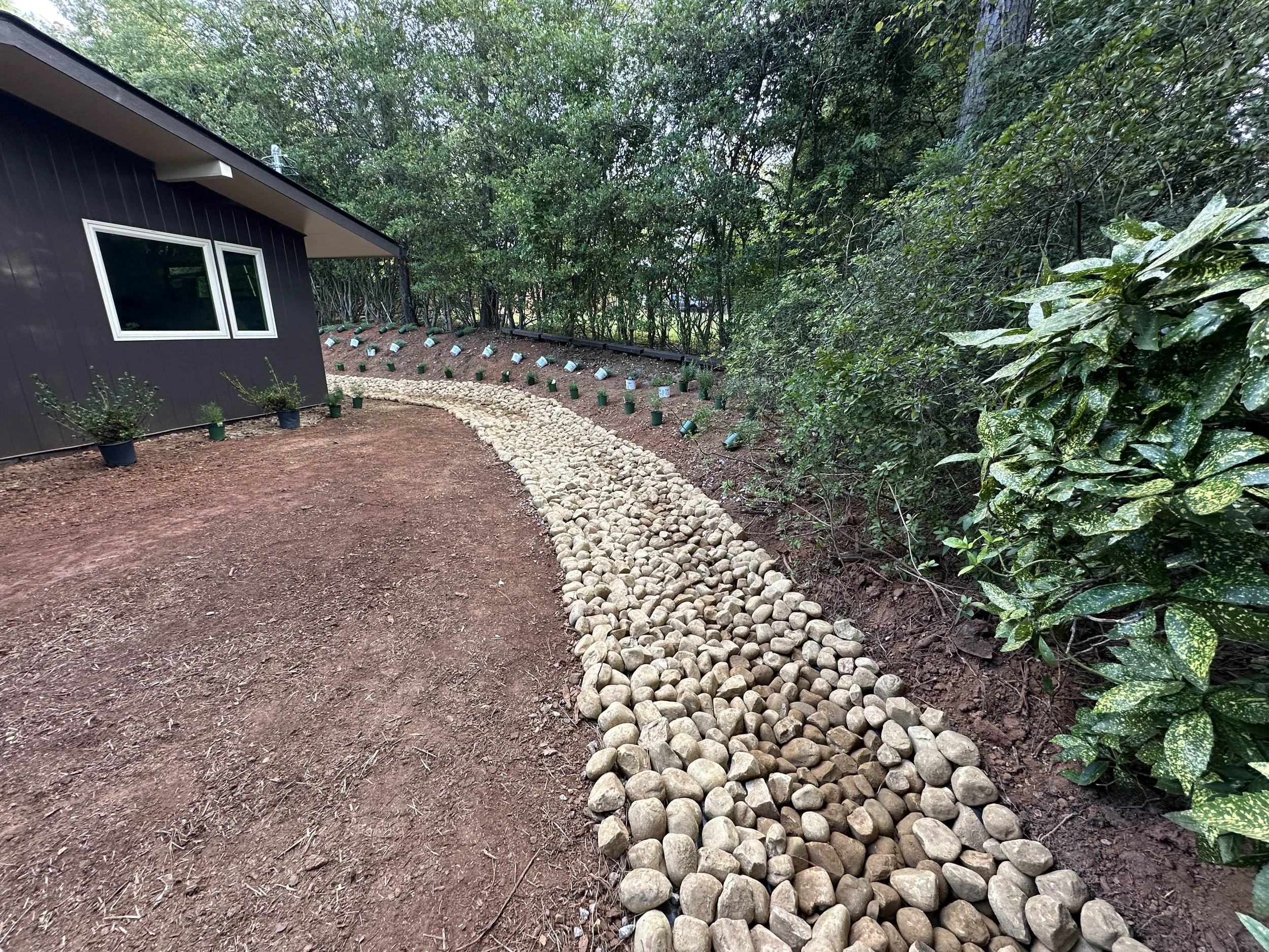 Long river rock drainage channel along home foundation through wooded backyard slope