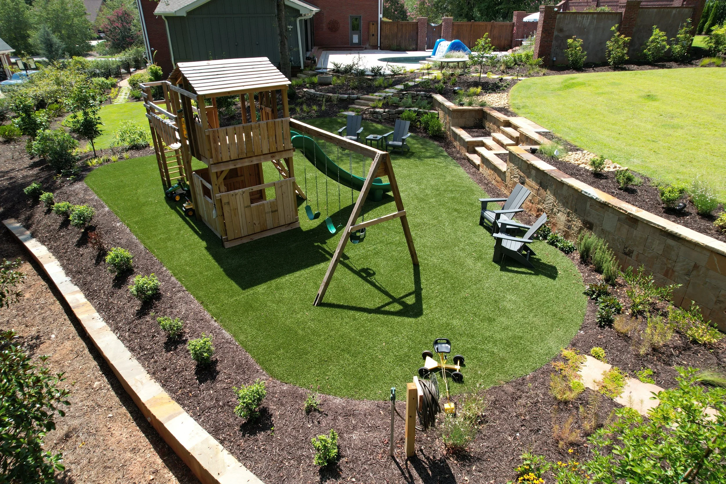 Artificial turf play area with children's swing set and play structure surrounded by stone retaining walls and mulch beds installed by Southern Grounds Landscaping in Athens Georgia