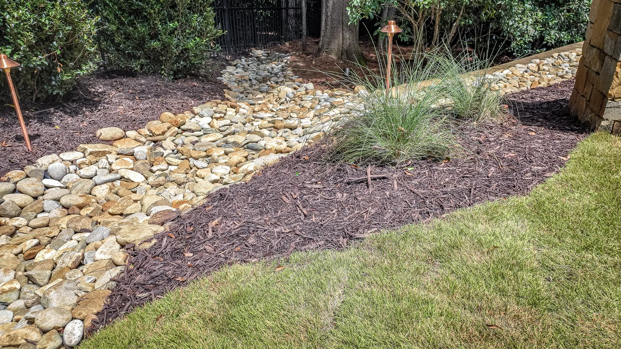 River rock drainage channel with landscape lighting fixture integrated into mulched garden bed