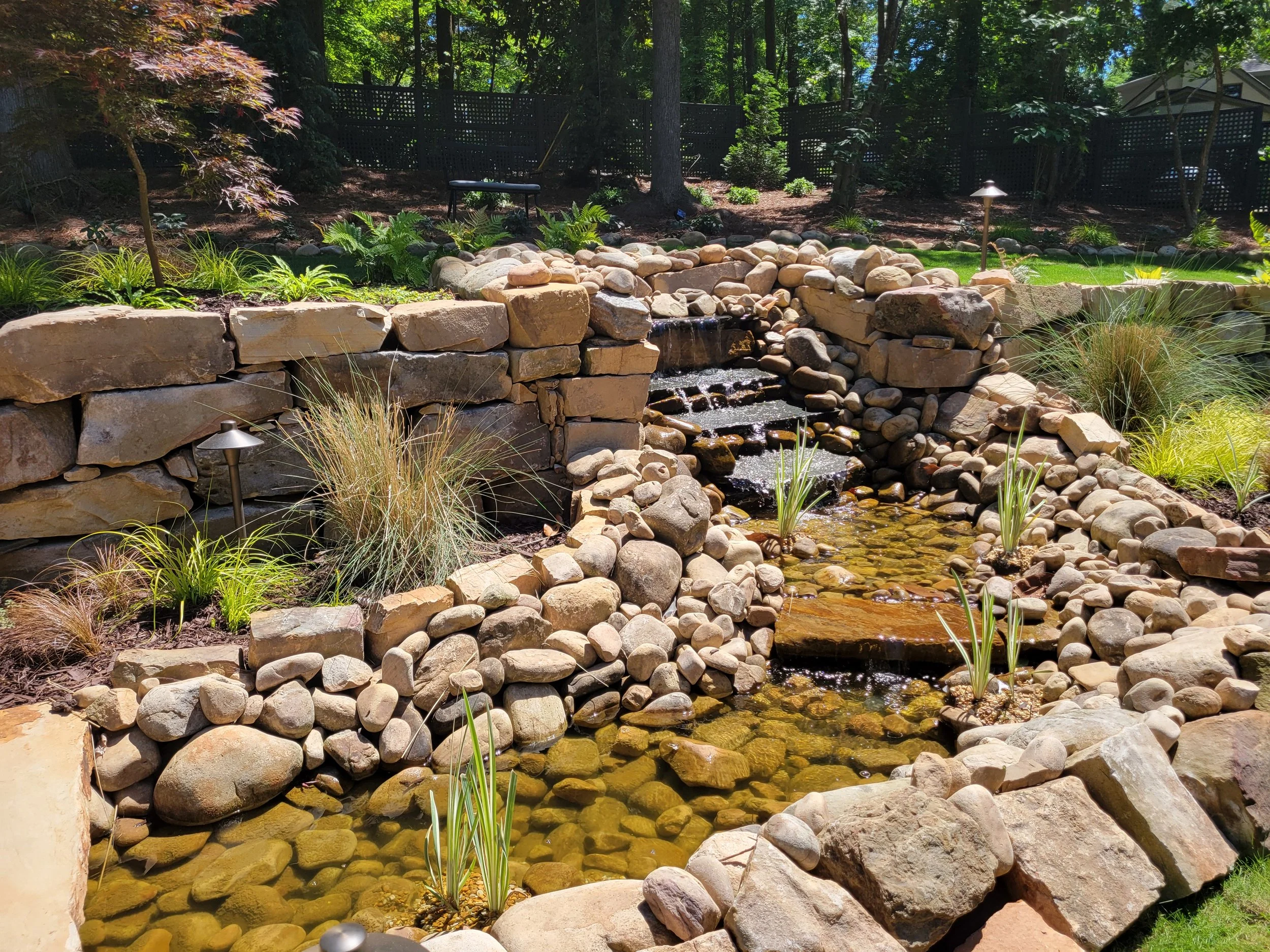 Natural stone pond with multi-tier waterfall, river rock borders, and landscape lighting accents.